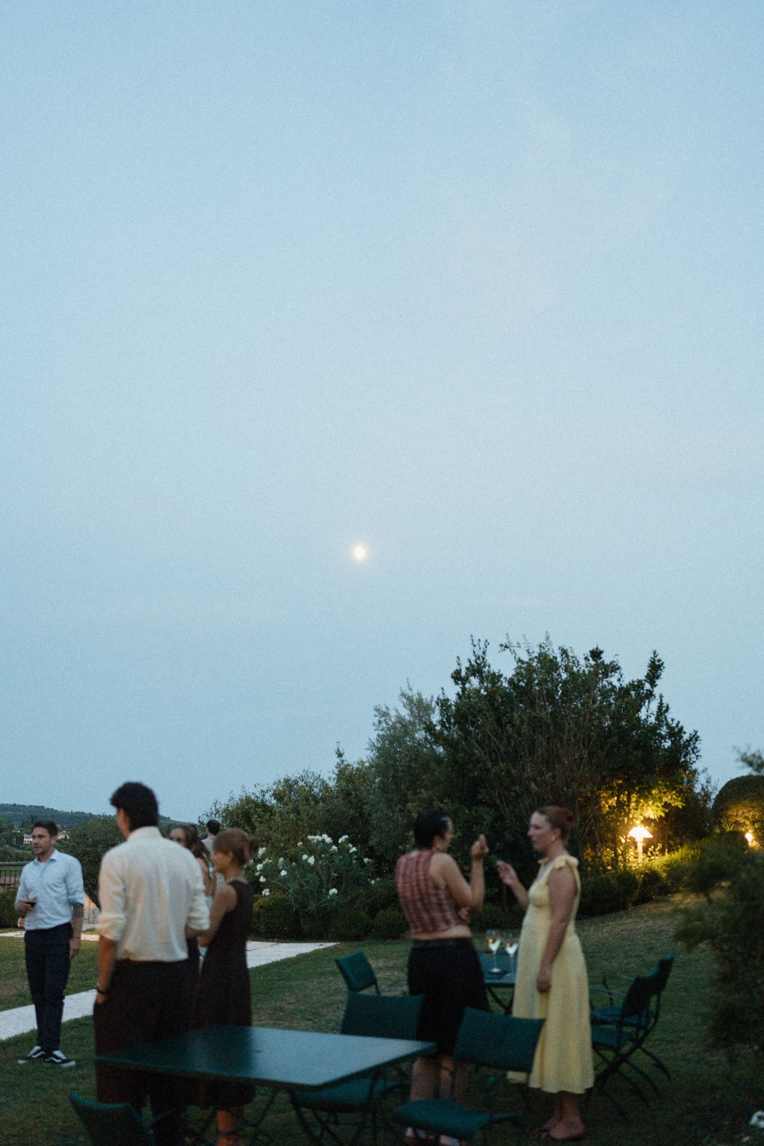 Guests mingle outdoors at dusk during an elegant garden party with moonrise visible in the twilight sky.