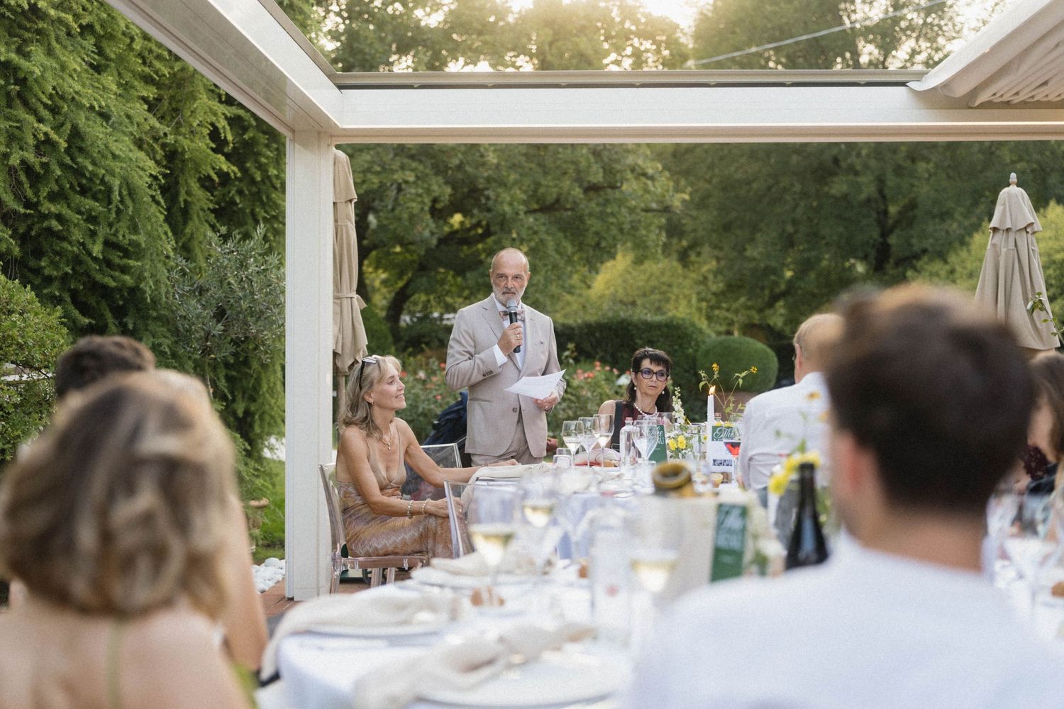 Man giving speech at elegant outdoor wedding reception dinner with guests seated at decorated tables.
