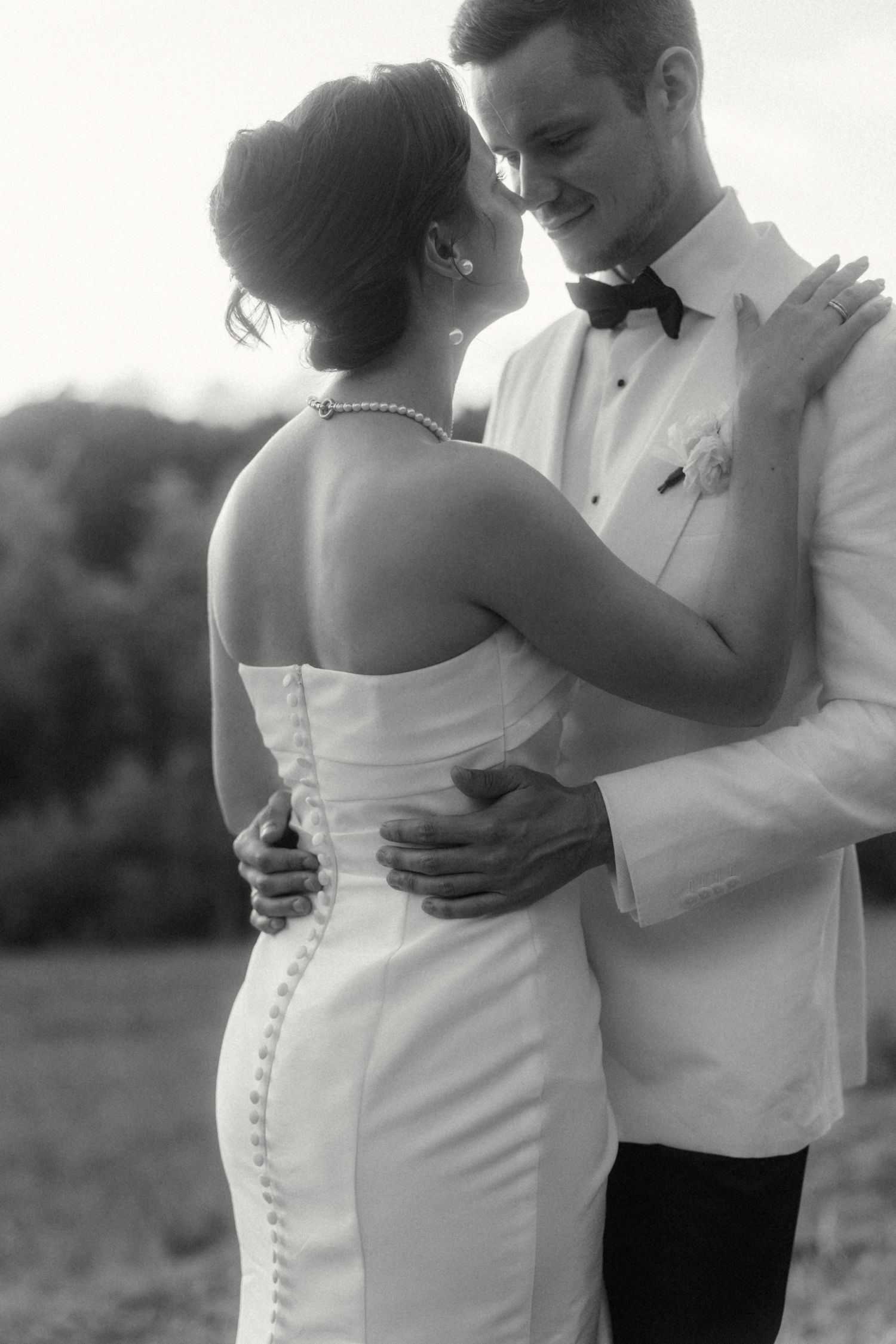 Black and white photo of bride and groom embracing outdoors, groom in white tuxedo with bow tie.