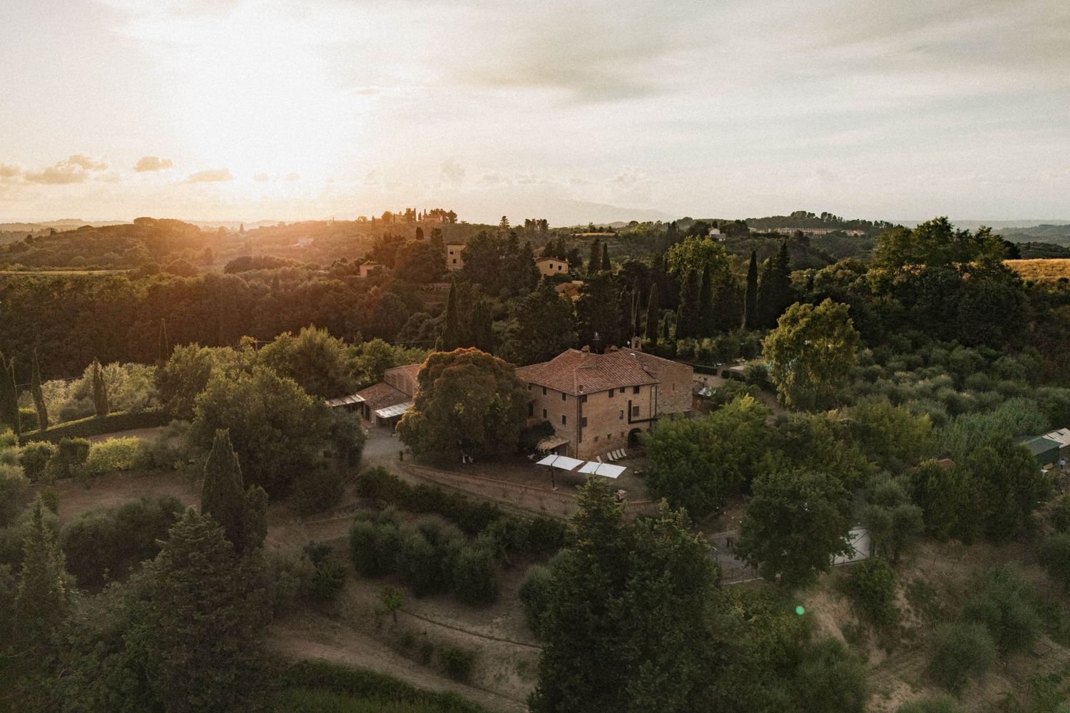 Aerial sunset view of rustic Tuscan countryside villa surrounded by cypress and olive trees.