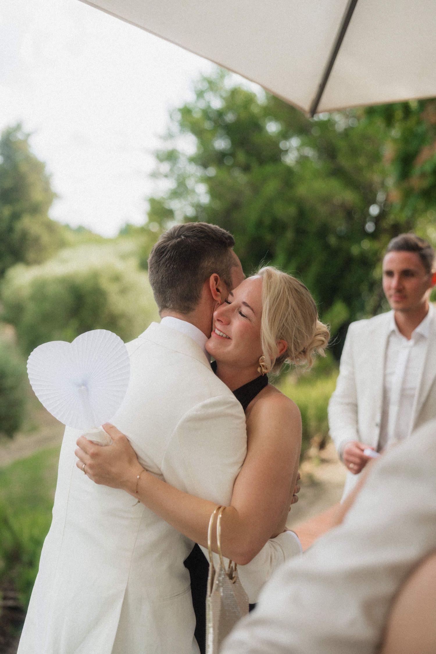 Bride and groom sharing joyful embrace during outdoor wedding ceremony holding heart fan.