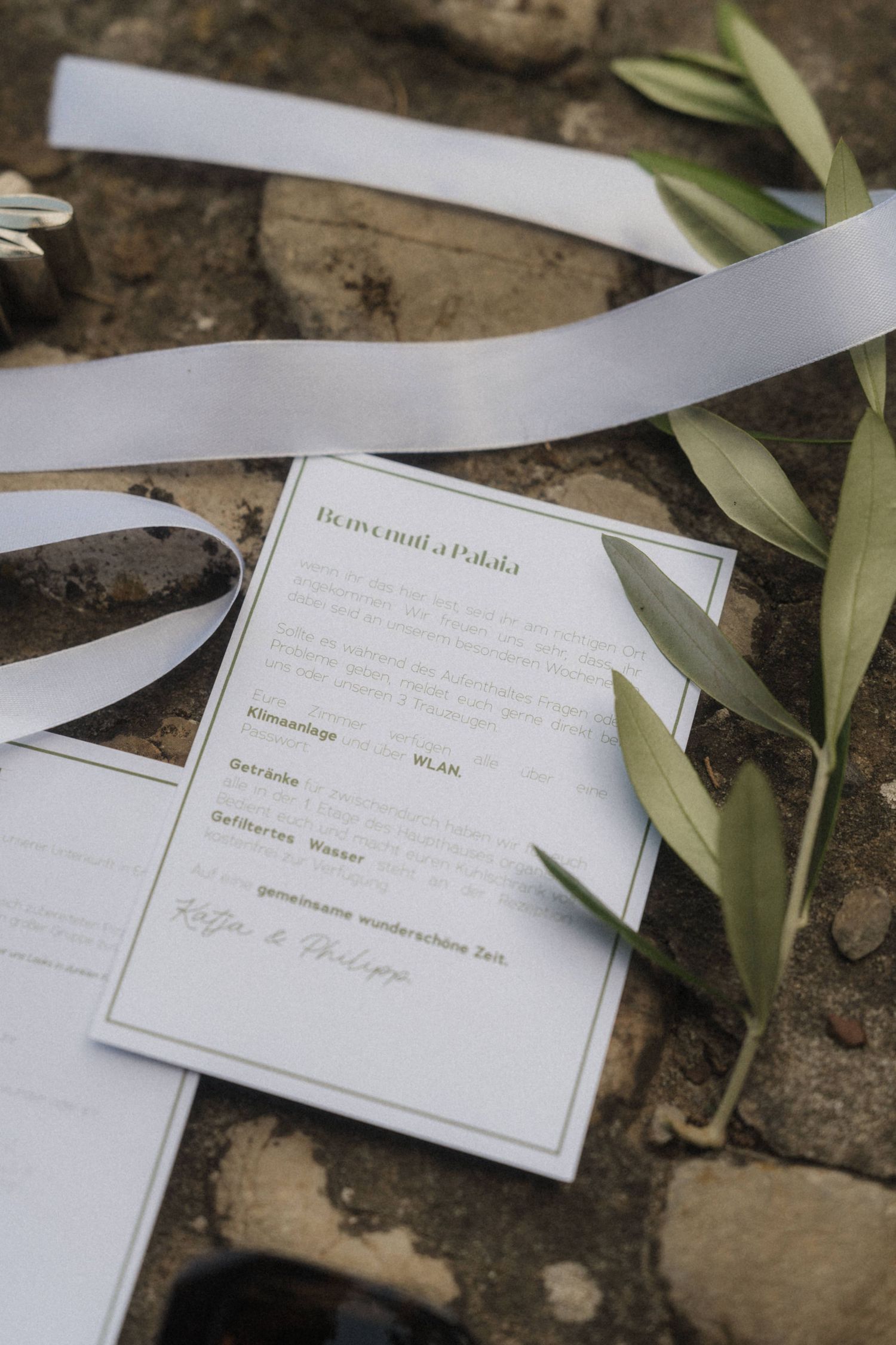 Elegant wedding stationery card with gold text reading Benvenuti a Palaia, surrounded by olive branches and white ribbon.