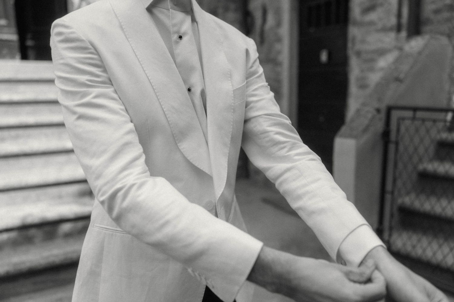 Elegant man in white tuxedo jacket adjusting cufflinks outdoors in black and white photo.
