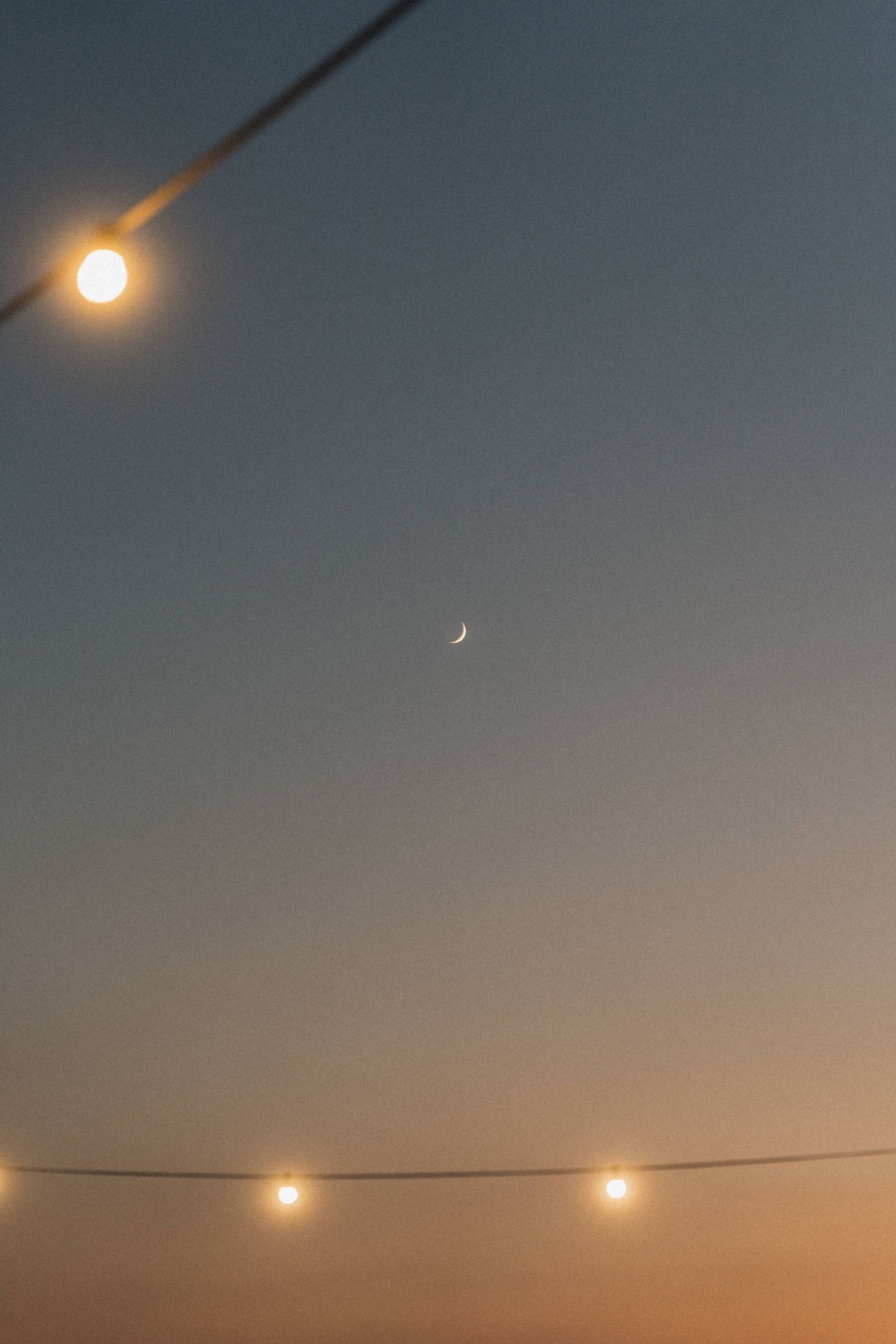 Crescent moon and string lights glowing against a warm dusk sky at outdoor wedding.