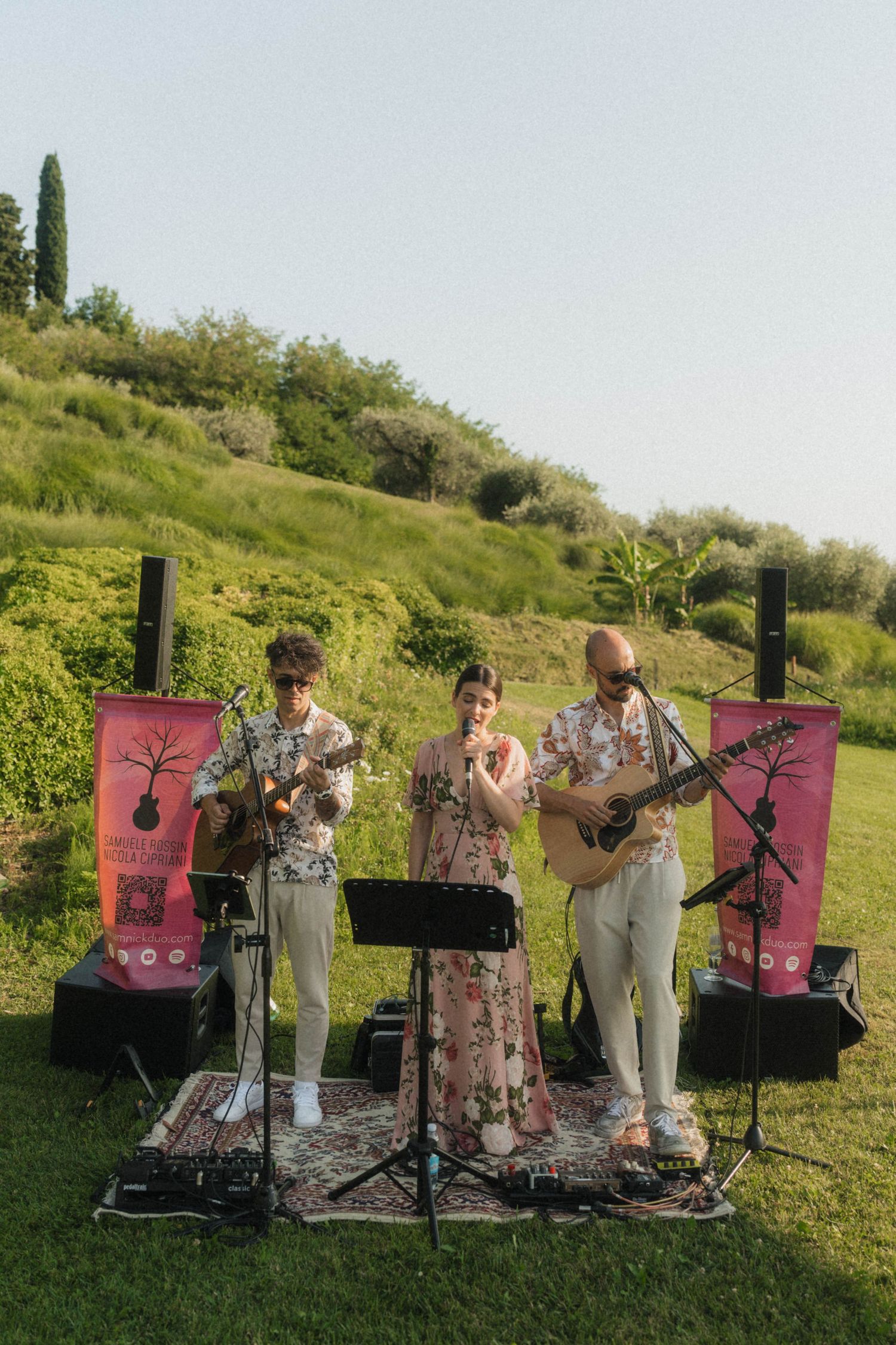 Three-piece live band performs outdoors at wedding reception on lush green hillside at dusk.