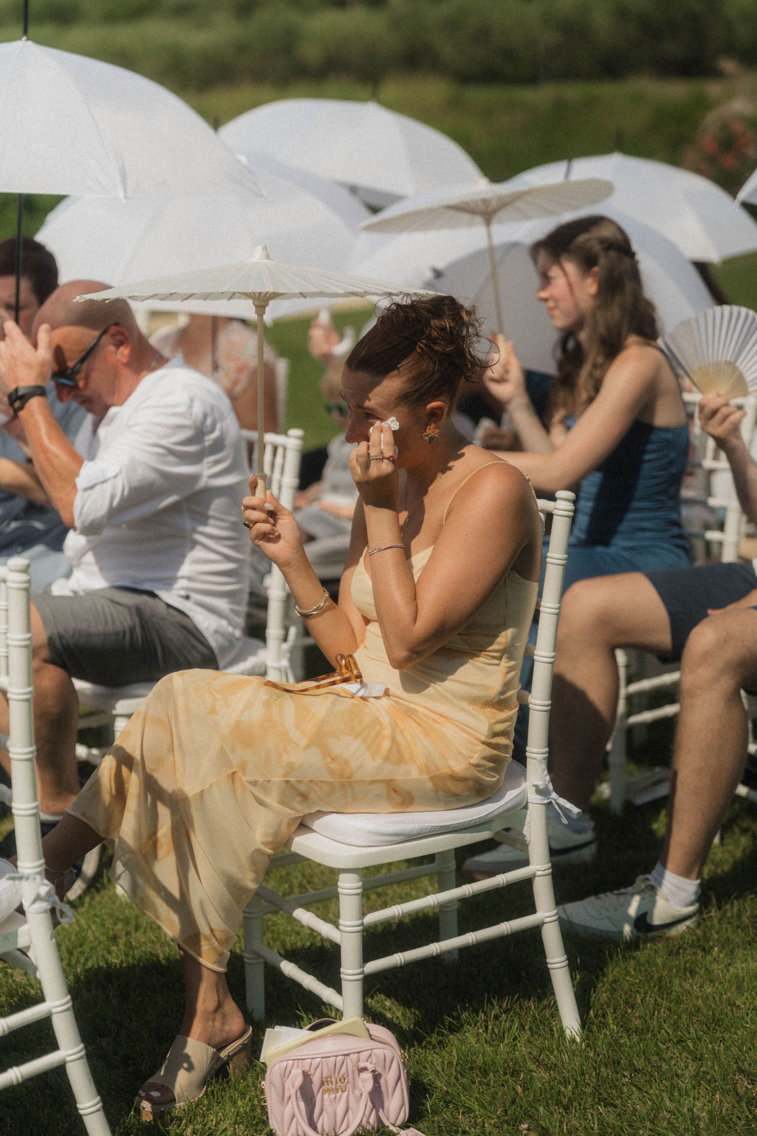 Wedding guest in yellow floral dress using hand fan and parasol at sunny outdoor ceremony.