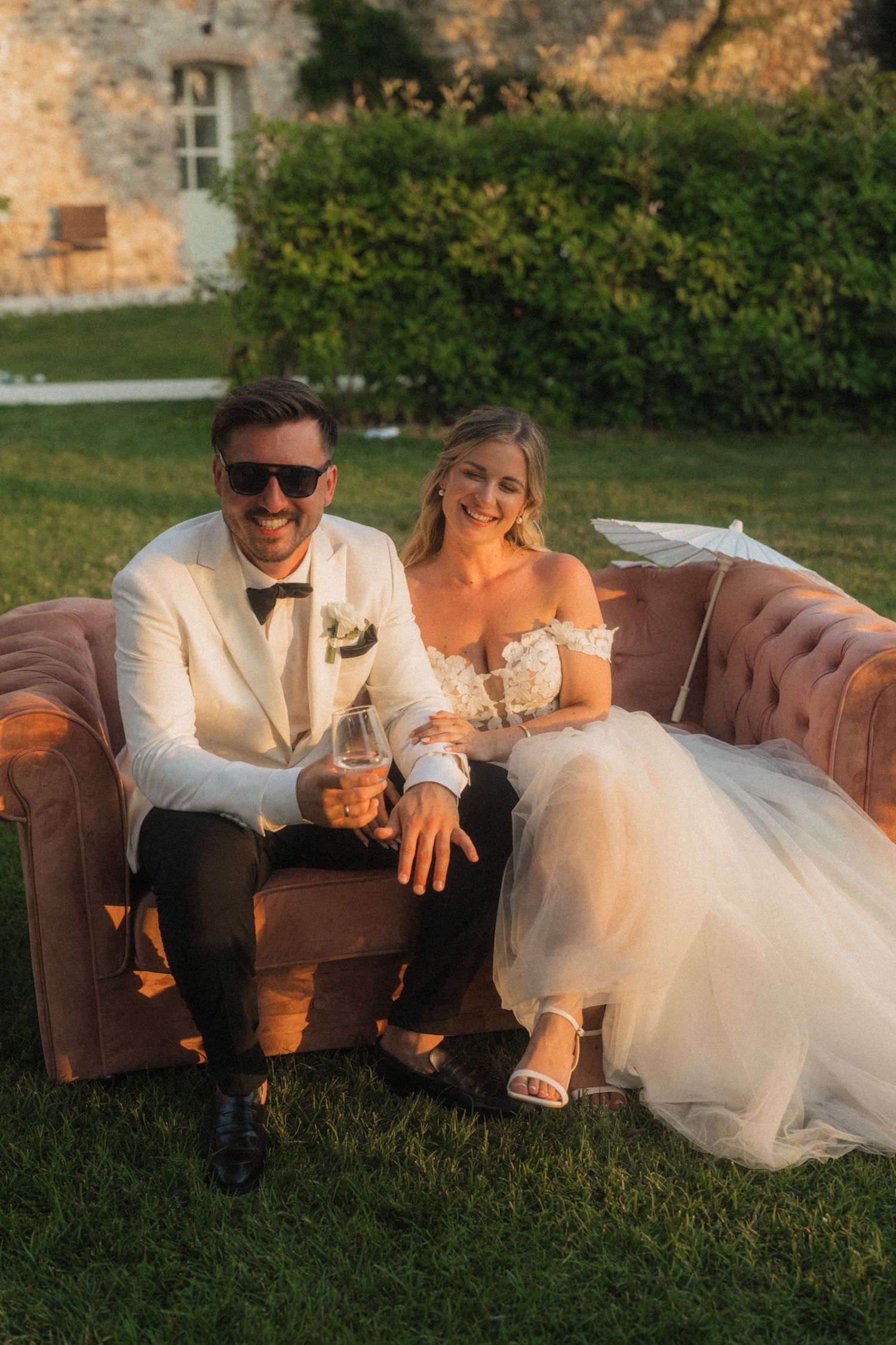 Bride and groom sitting on velvet sofa outdoors at golden hour.