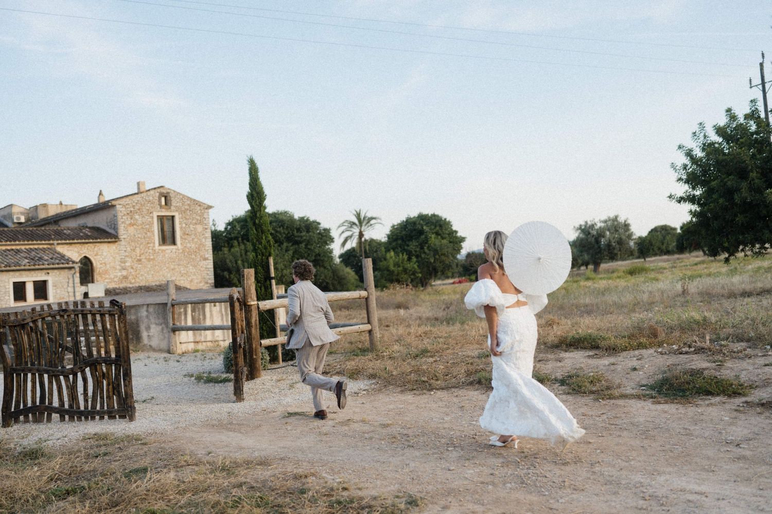 Bride and groom running playfully with a large balloon on a rustic dirt path near a stone farmhouse.