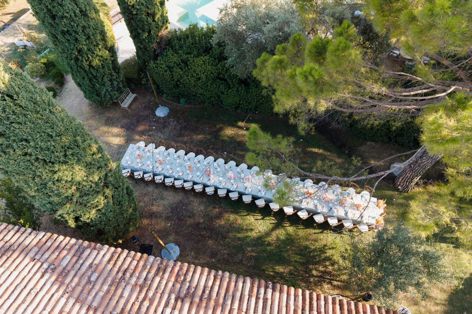 Aerial view of a long elegant outdoor dining table setup surrounded by lush greenery and cypress trees.