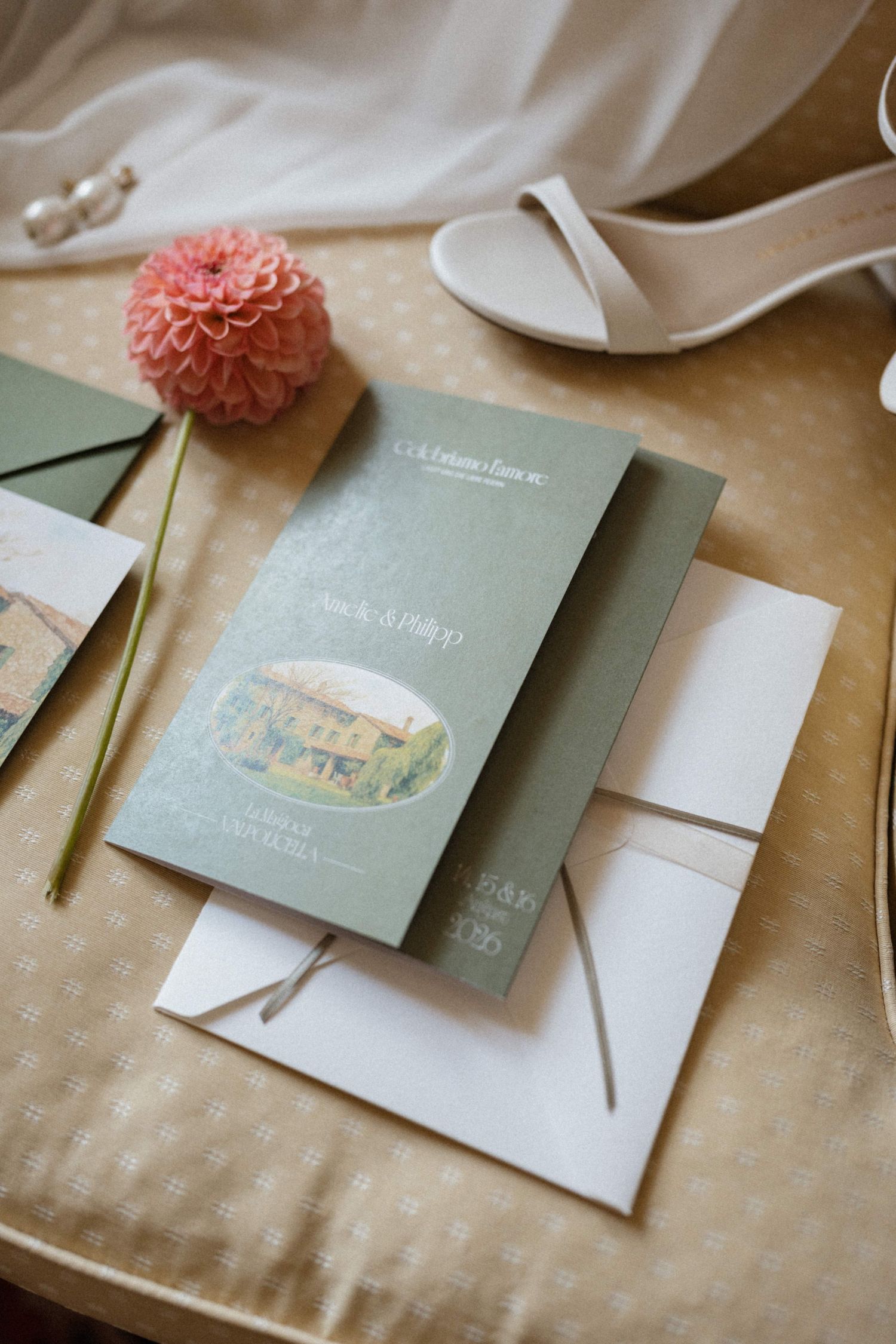Elegant sage green wedding invitation suite with coral dahlia, white envelope, and bridal heels on golden surface.
