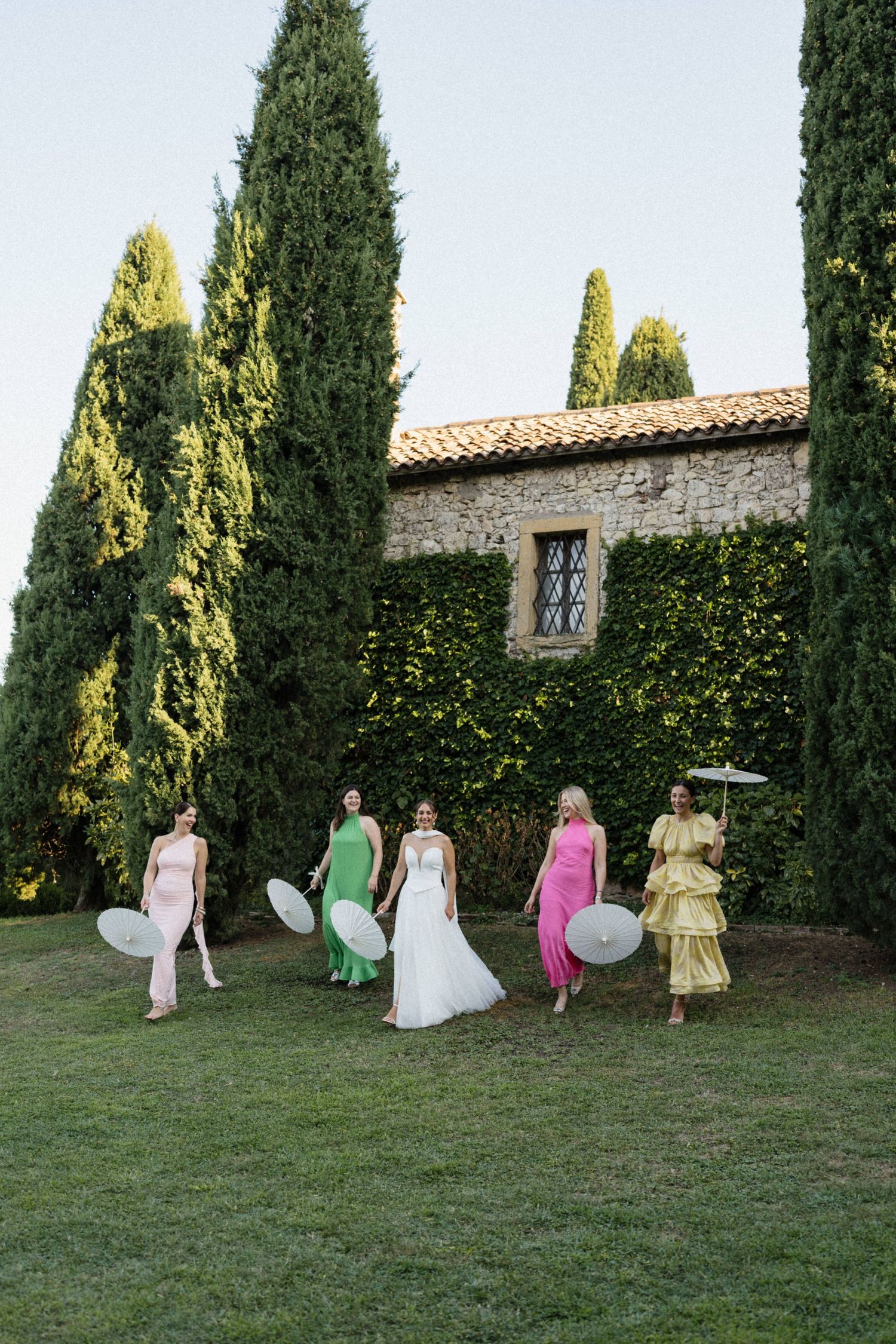 Bride and bridesmaids in colorful gowns walk with parasols past tall cypress trees at a Tuscan stone villa.