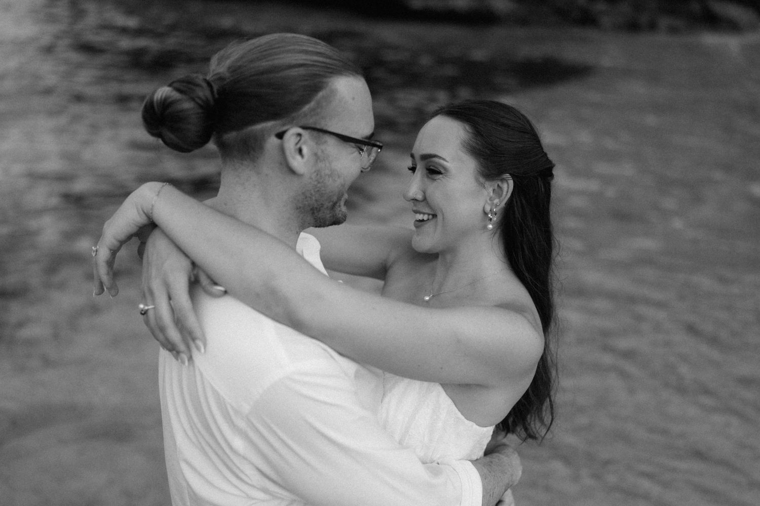 Black and white photo of a smiling couple embracing closely by the water, the man in a white shirt with a bun and glasses.
