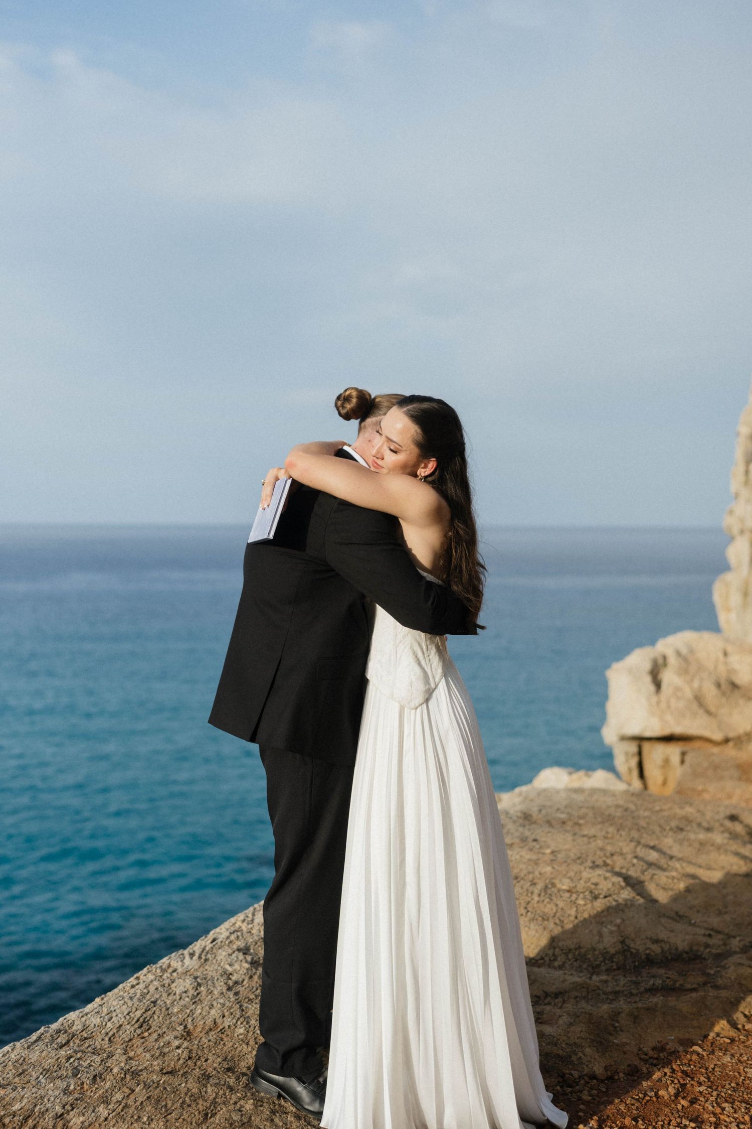 Bride and groom share a heartfelt embrace on a rocky cliff overlooking the deep blue Mediterranean sea.