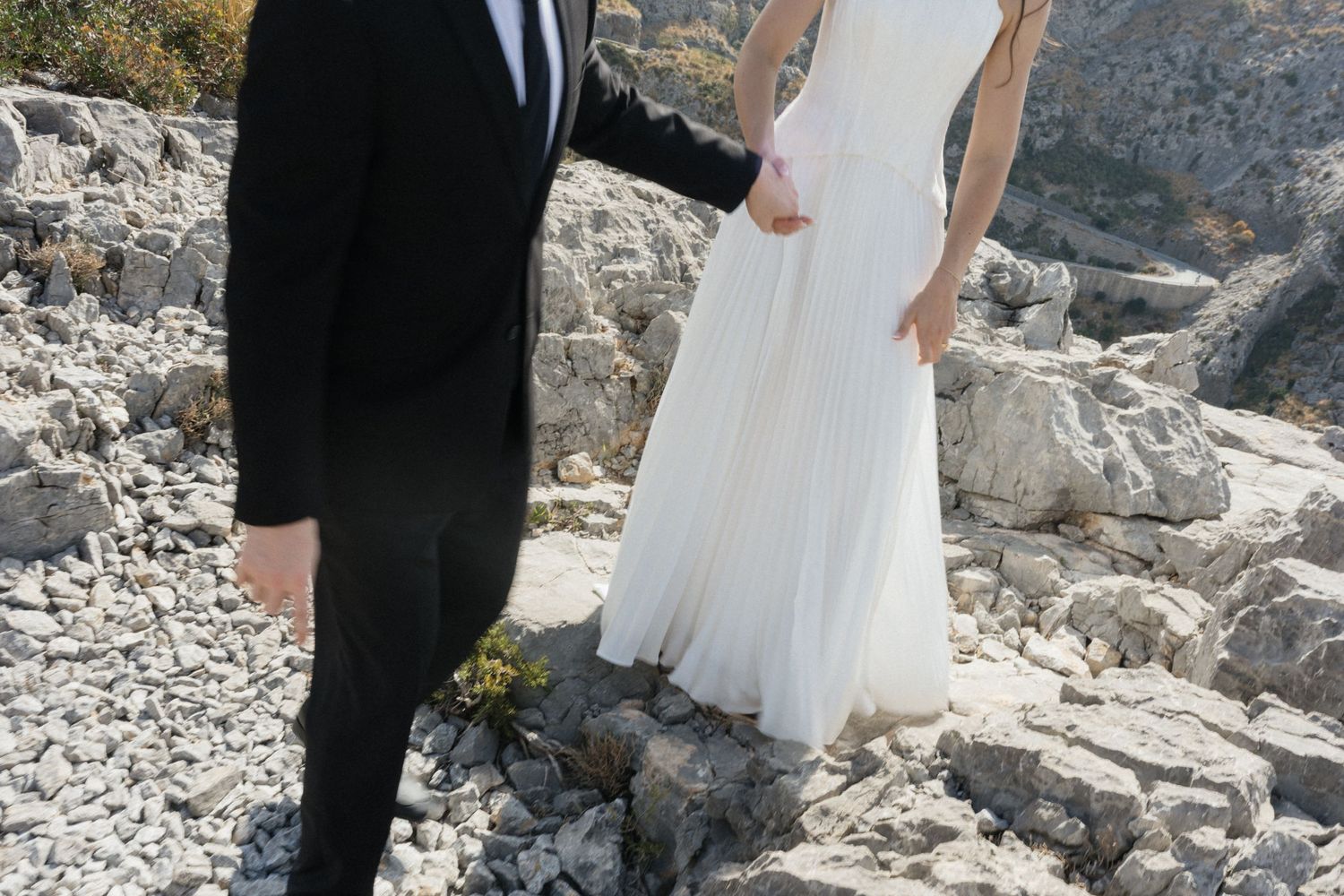 Bride in a flowing white gown holds hands with a groom in a dark suit while standing on rugged rocky terrain at an outdoor wedding.