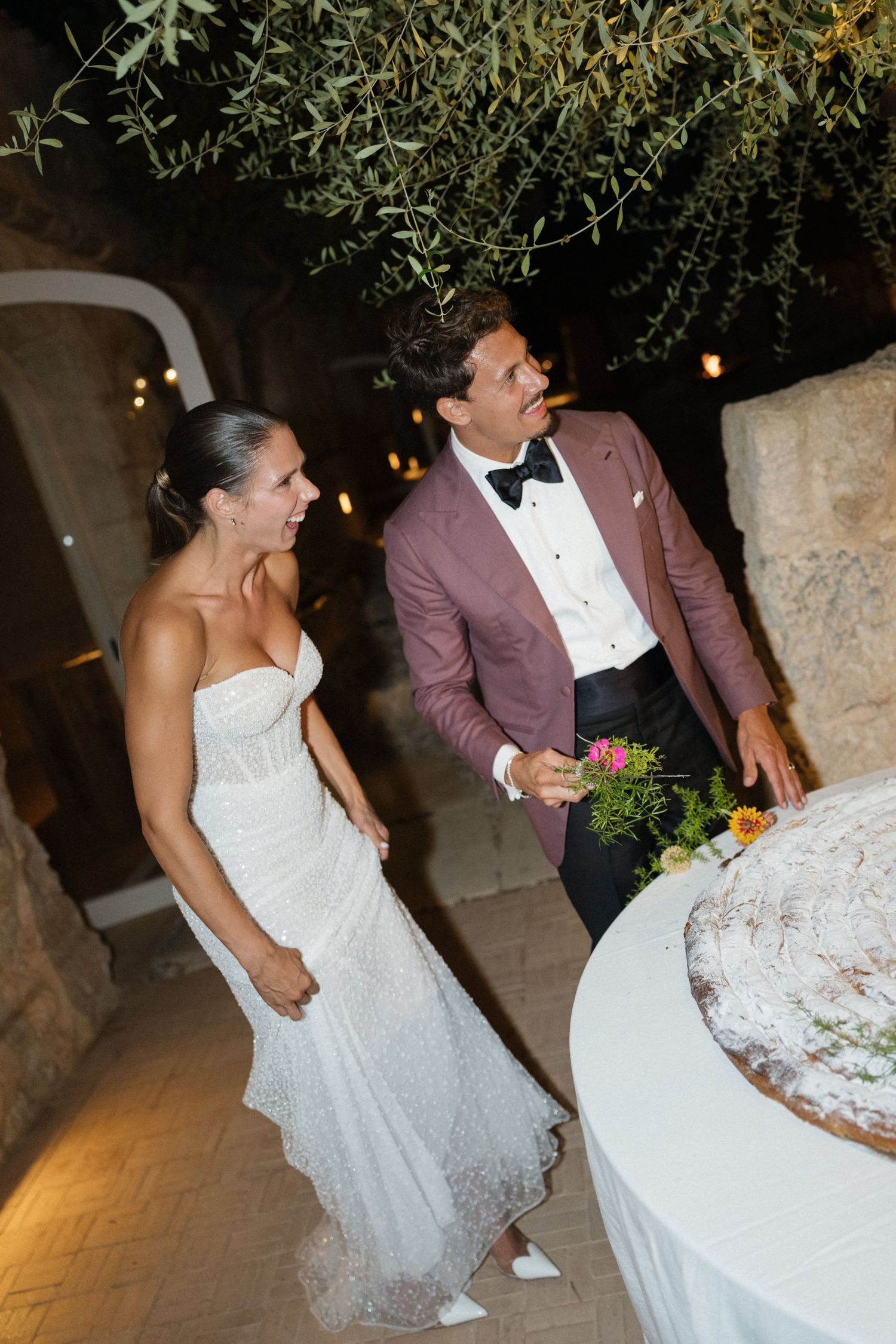 A bride in a sparkling strapless gown and groom in a burgundy jacket and bow tie smile beside their wedding cake at night.