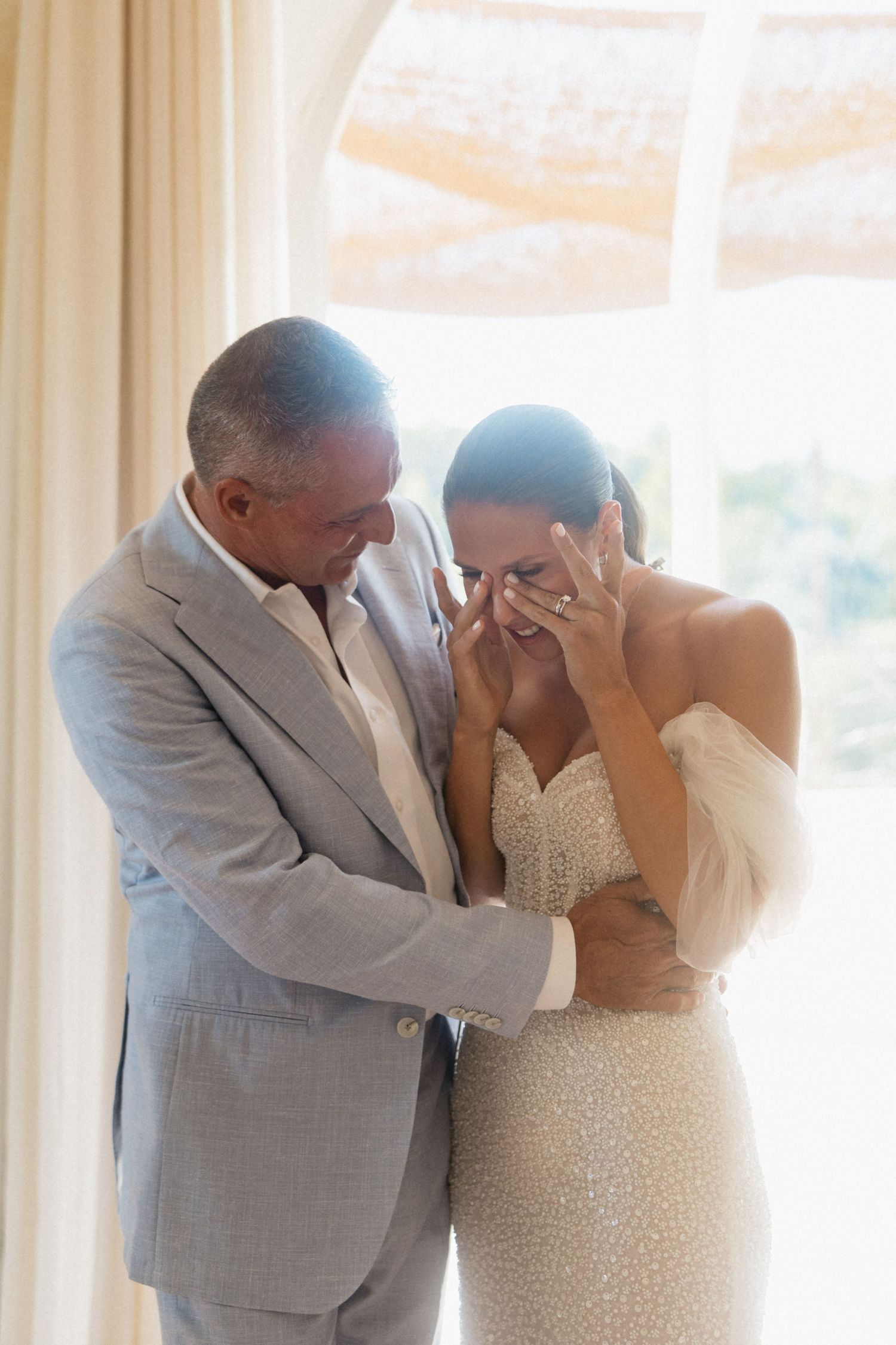 Emotional bride covering her face with her hands as her smiling father in a gray suit embraces her in a sunlit room.