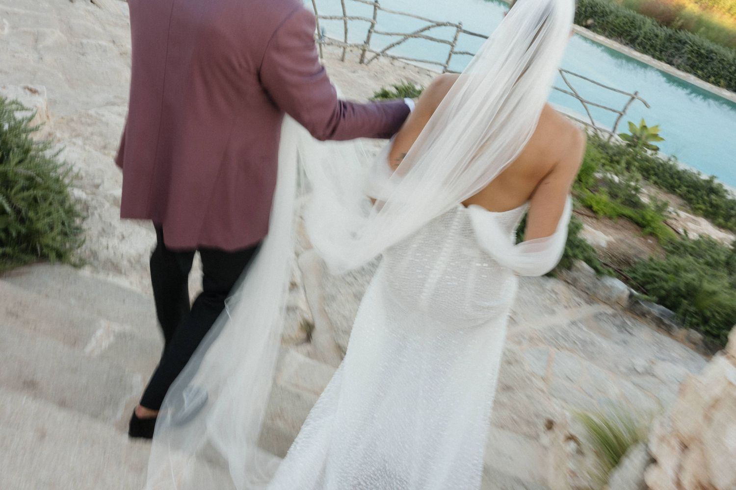 Bride in a white gown with a long flowing veil walks down stone steps beside a groom in a burgundy jacket near the sea.