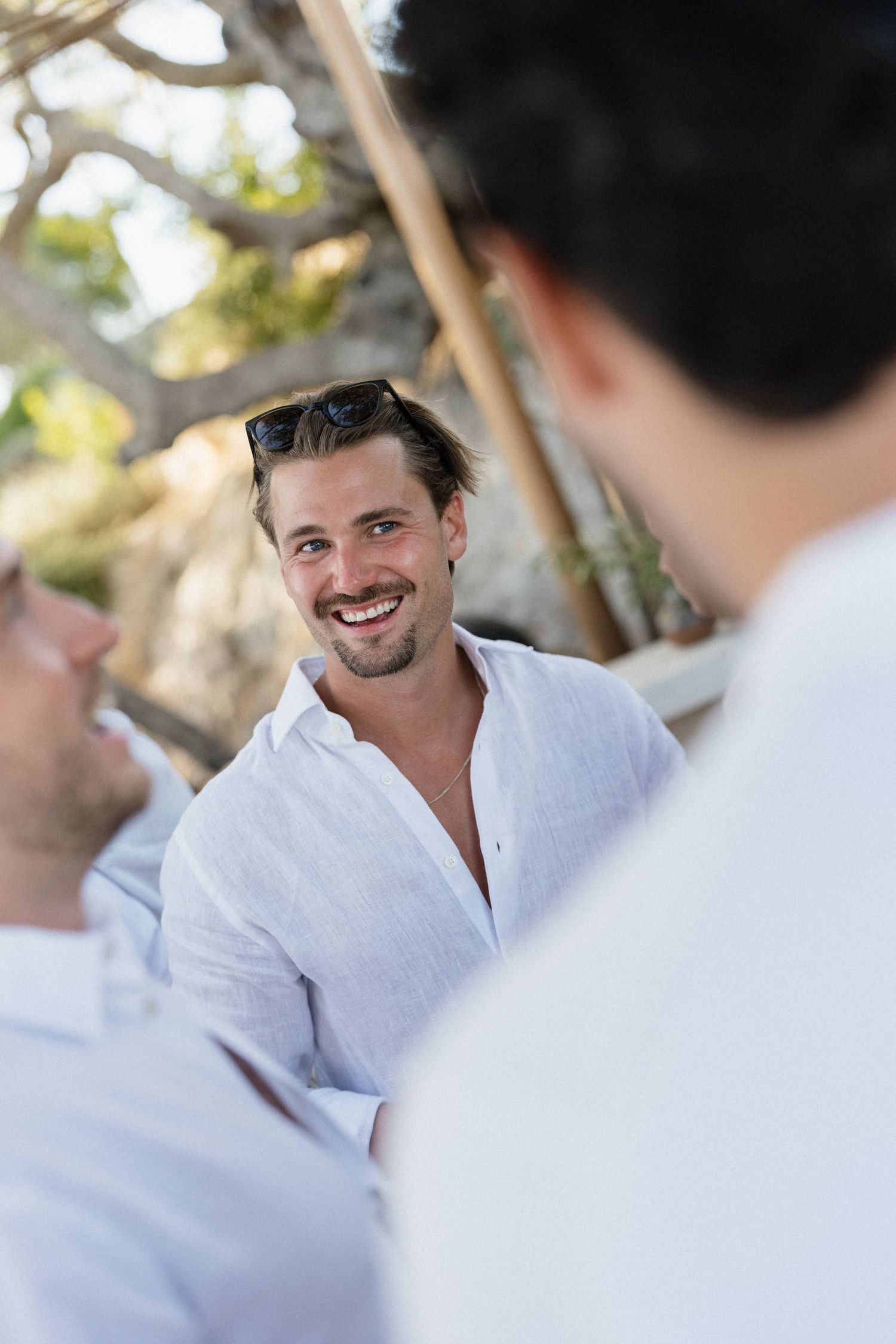 Smiling man in a white linen shirt with sunglasses on his head chats with friends outdoors under sunlit trees.
