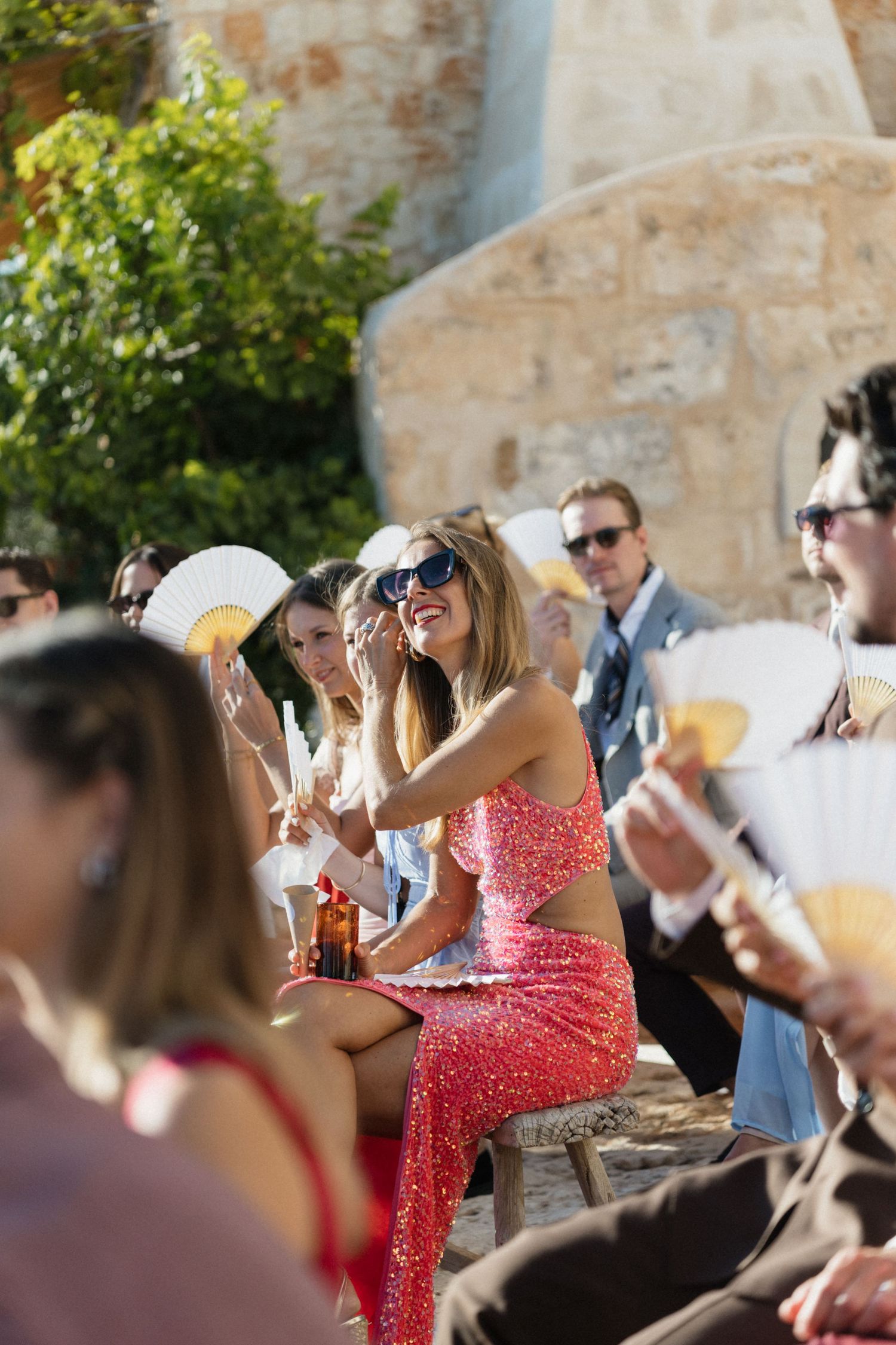A smiling woman in a sparkling coral sequin dress and sunglasses sits among wedding guests holding straw hand fans at an outdoor ceremony.