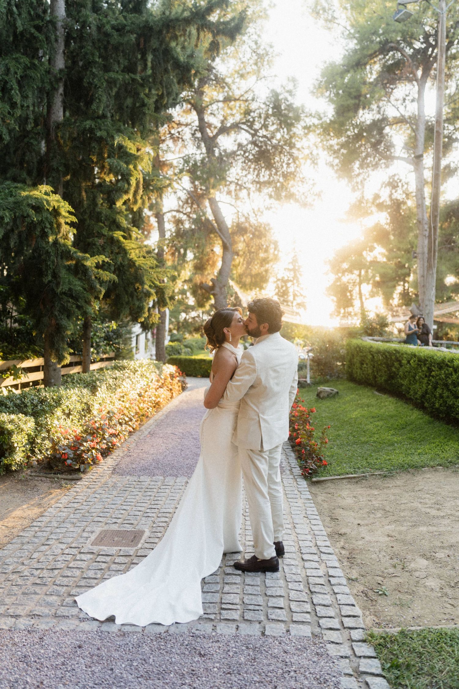 Bride and groom sharing a kiss on a cobblestone path surrounded by lush greenery and golden sunset light.