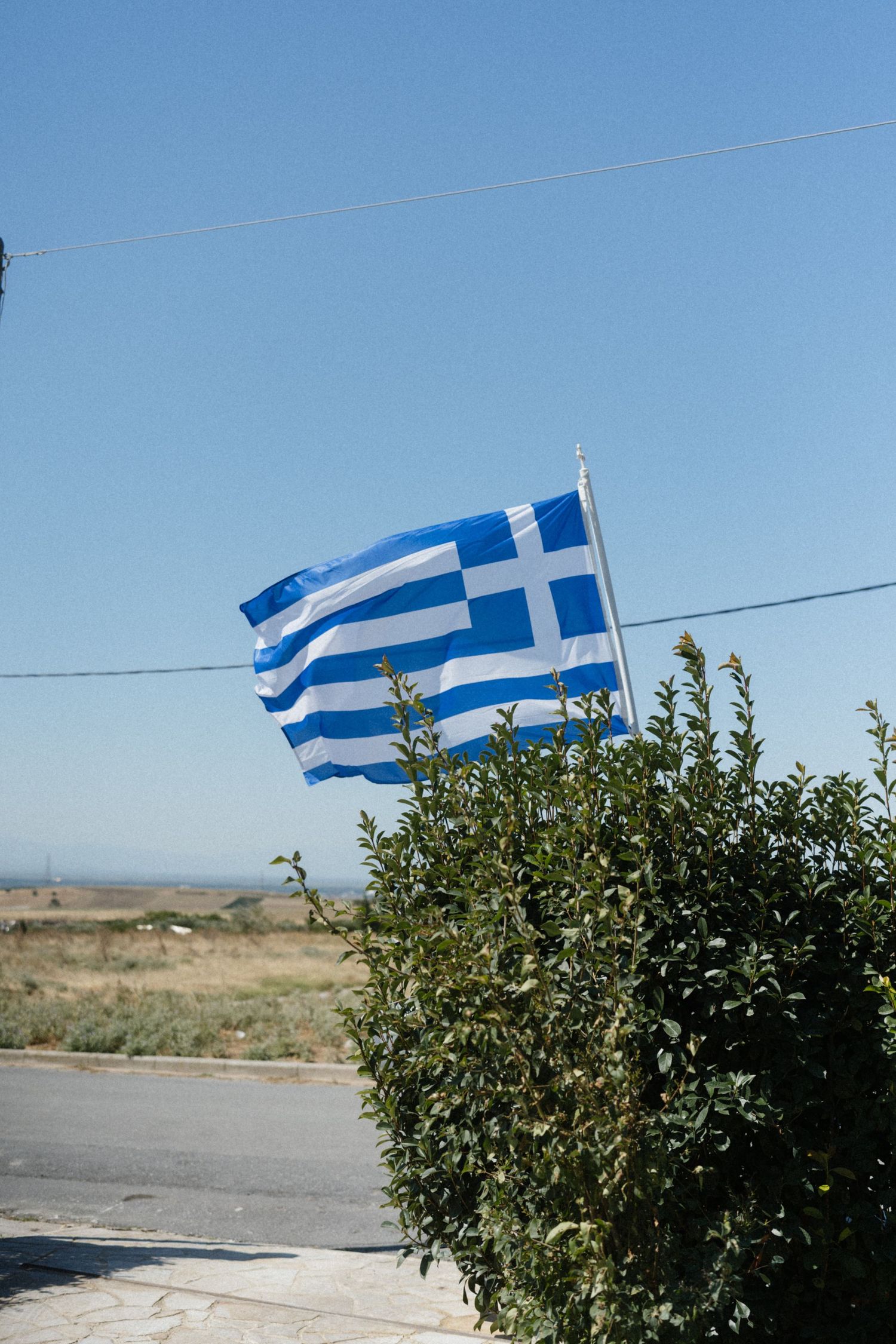 Greek flag waving in the breeze against a clear blue sky, with green shrubs and a rural landscape below.