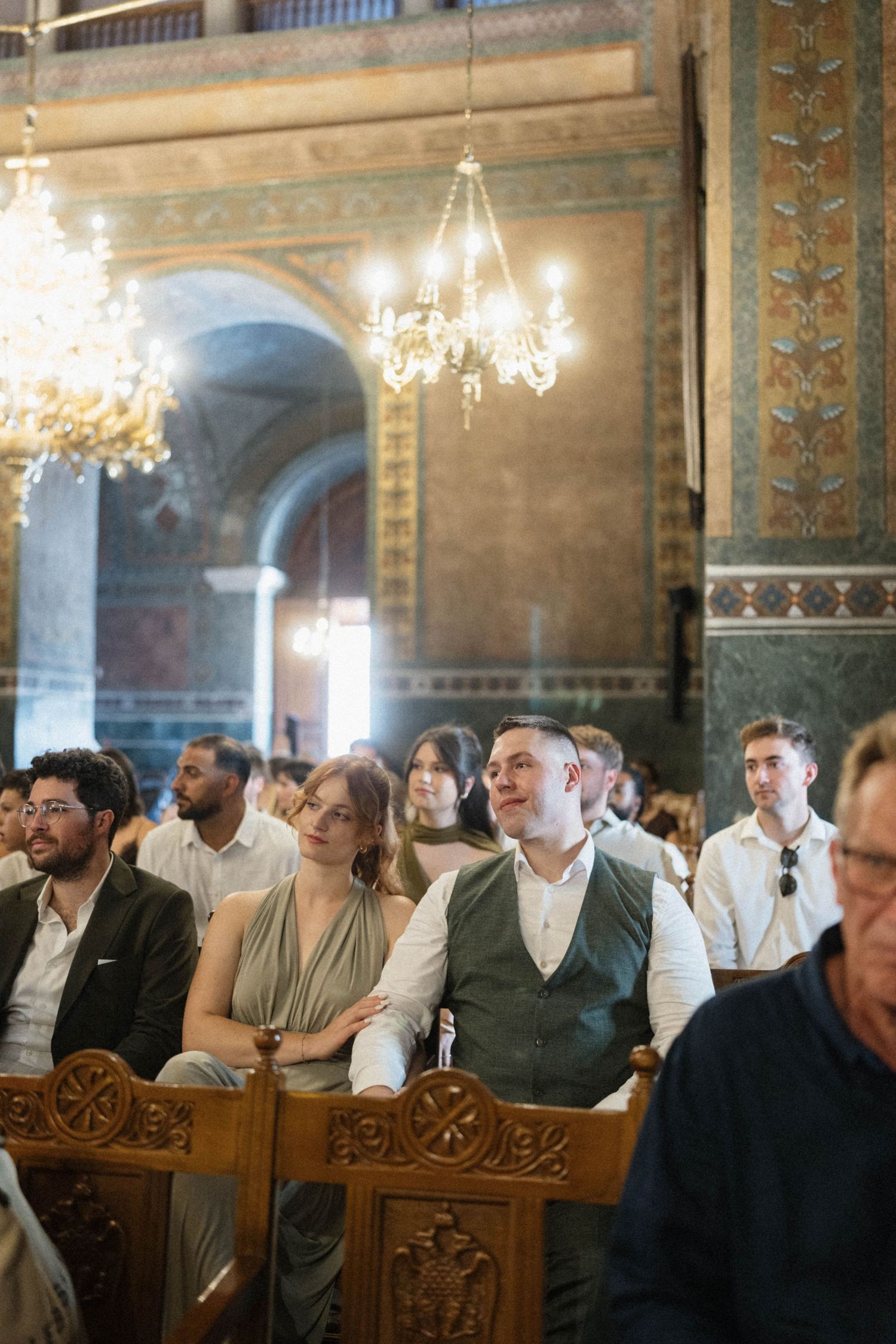 Wedding guests seated in ornate church pews beneath crystal chandeliers during a elegant ceremony.