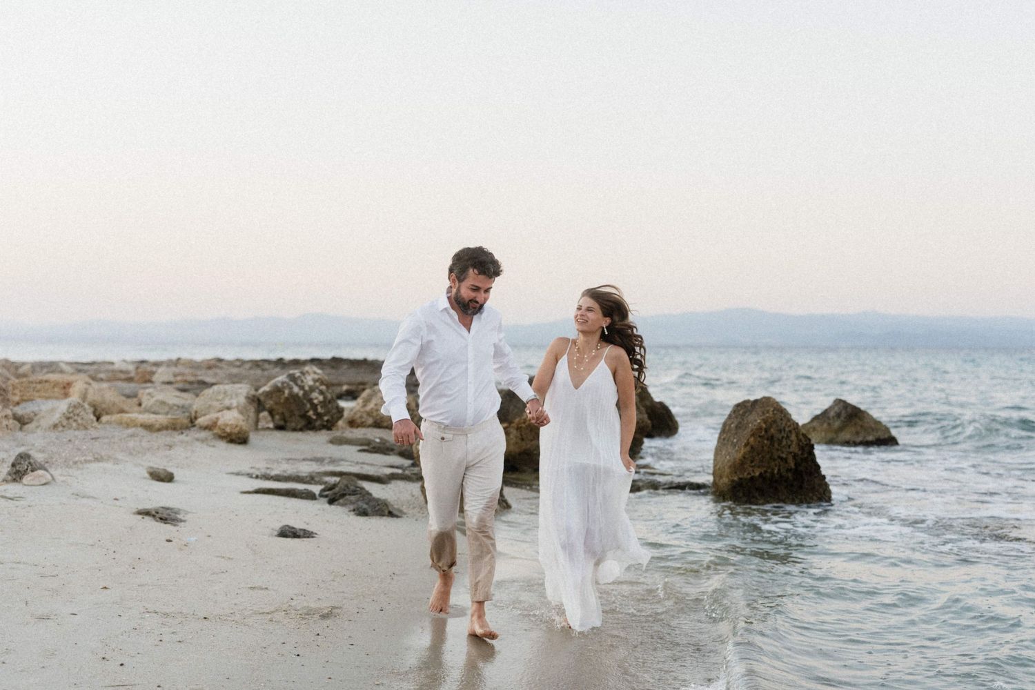Couple holding hands and walking barefoot along a sandy beach shore in white wedding attire.