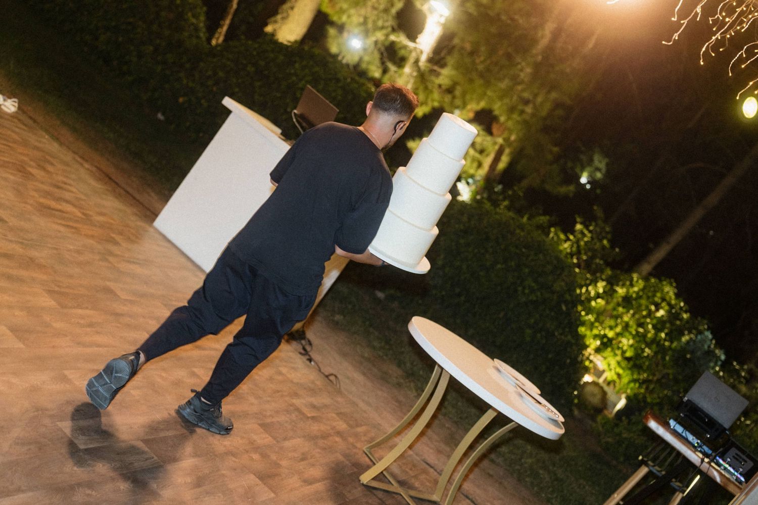 Man carrying large white cylindrical objects at an outdoor evening event with garden lighting.