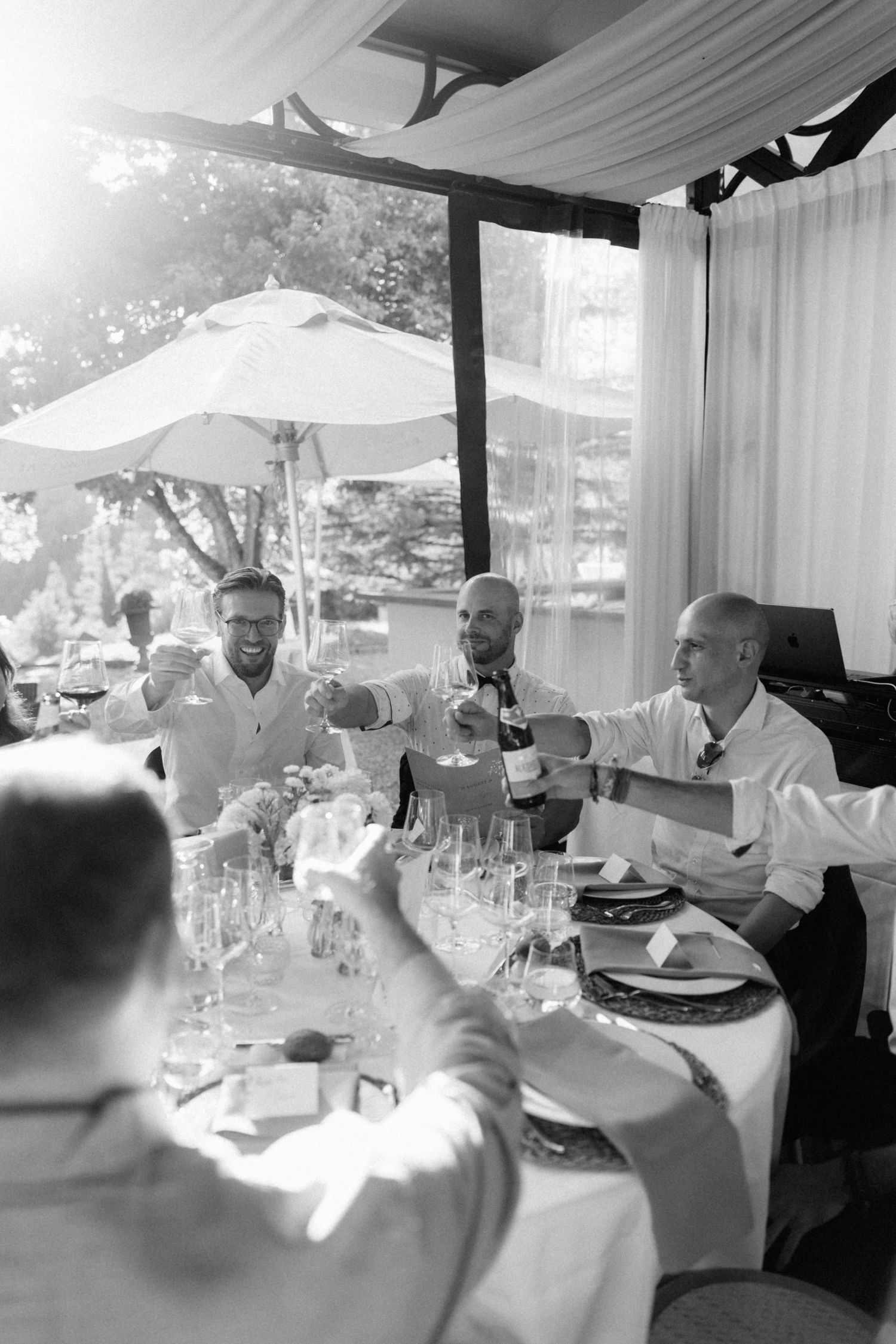 Black and white photo of groomsmen in white shirts toasting with a wine bottle at a sunlit wedding reception table.