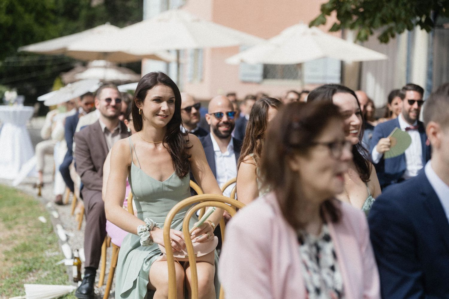 Wedding guests seated outdoors in summer attire, smiling and watching a ceremony at a scenic venue.