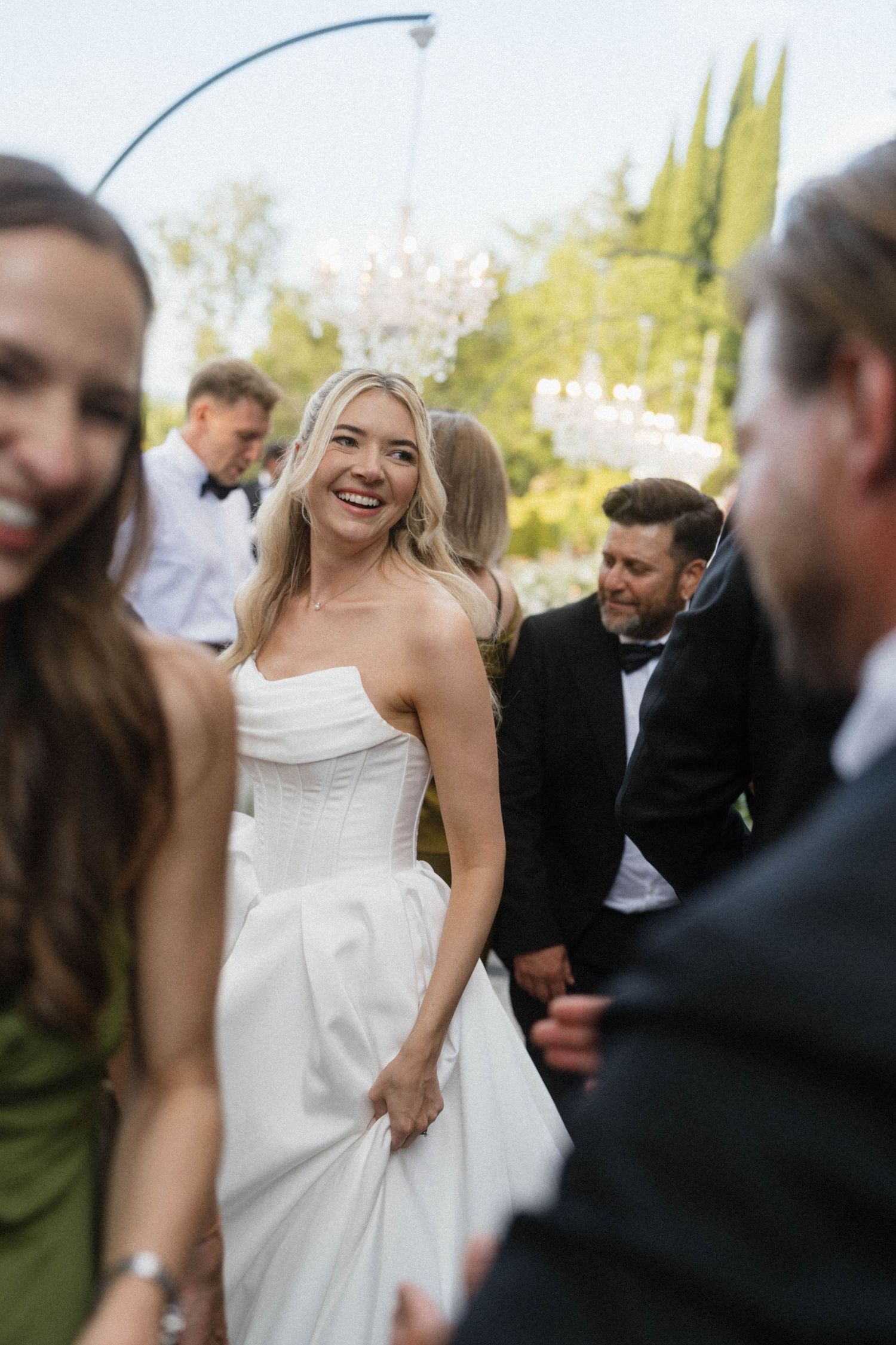 Radiant bride in strapless white gown smiling warmly at outdoor wedding reception with crystal chandelier.