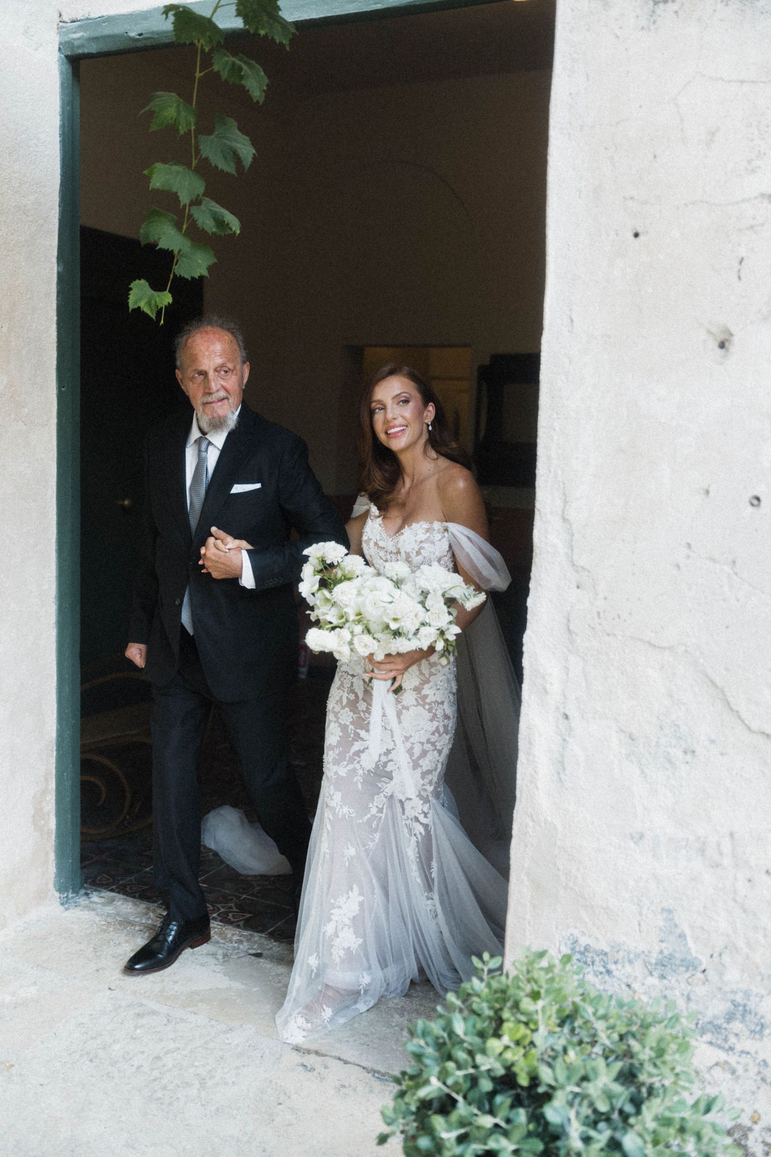Bride in lace gown holding white bouquet walks with escort through stone doorway.
