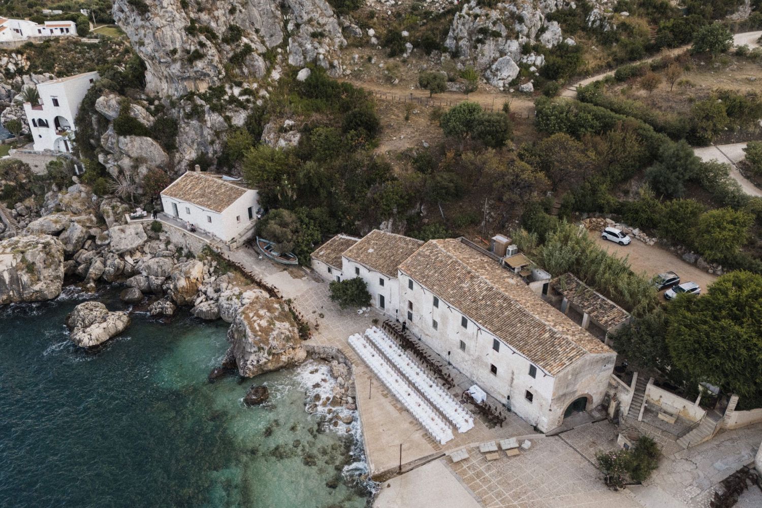 Aerial drone view of a historic stone building beside crystal clear turquoise water on rocky coastline.