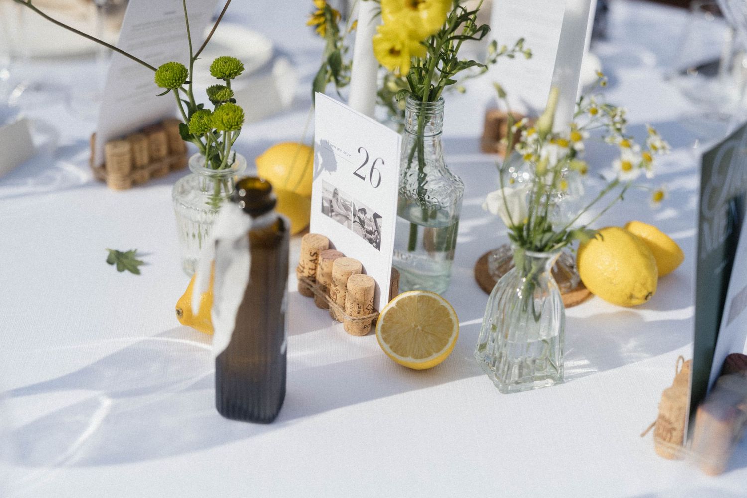 Wedding table decor with yellow flowers, lemons, glass bottles, and a table number card on a white tablecloth.
