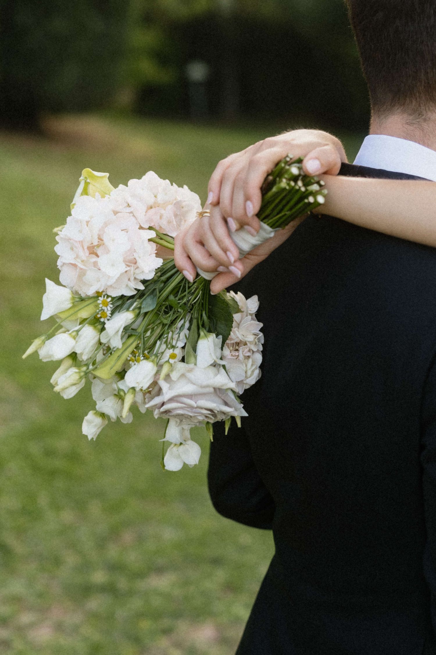 Bride holding a delicate white bridal bouquet of peonies and roses while embracing the groom in a black suit.
