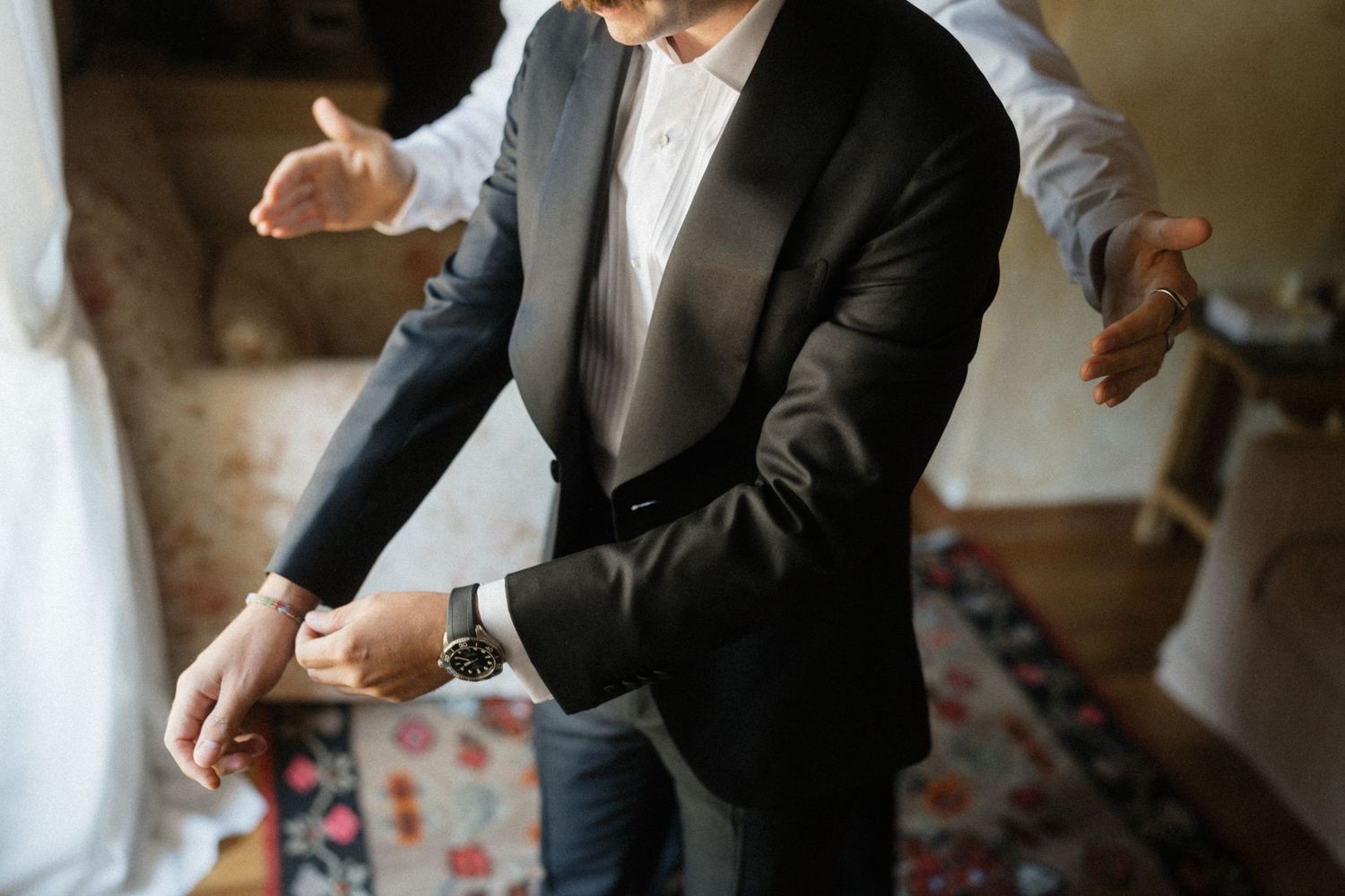 Groom in black tuxedo jacket checking his watch while being assisted during wedding prep.