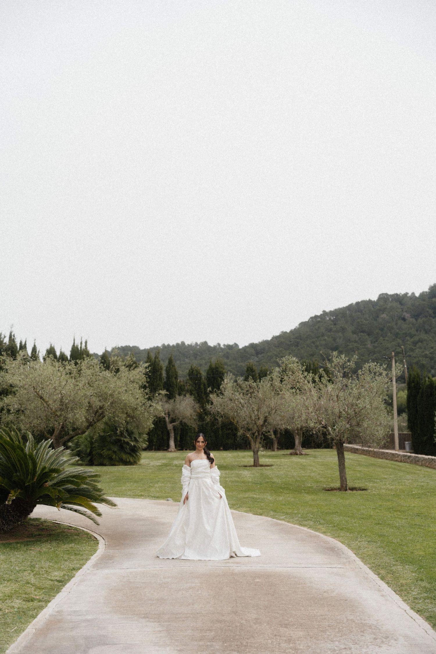 Elegant bride in flowing white off-shoulder gown walking on garden path with green hills.