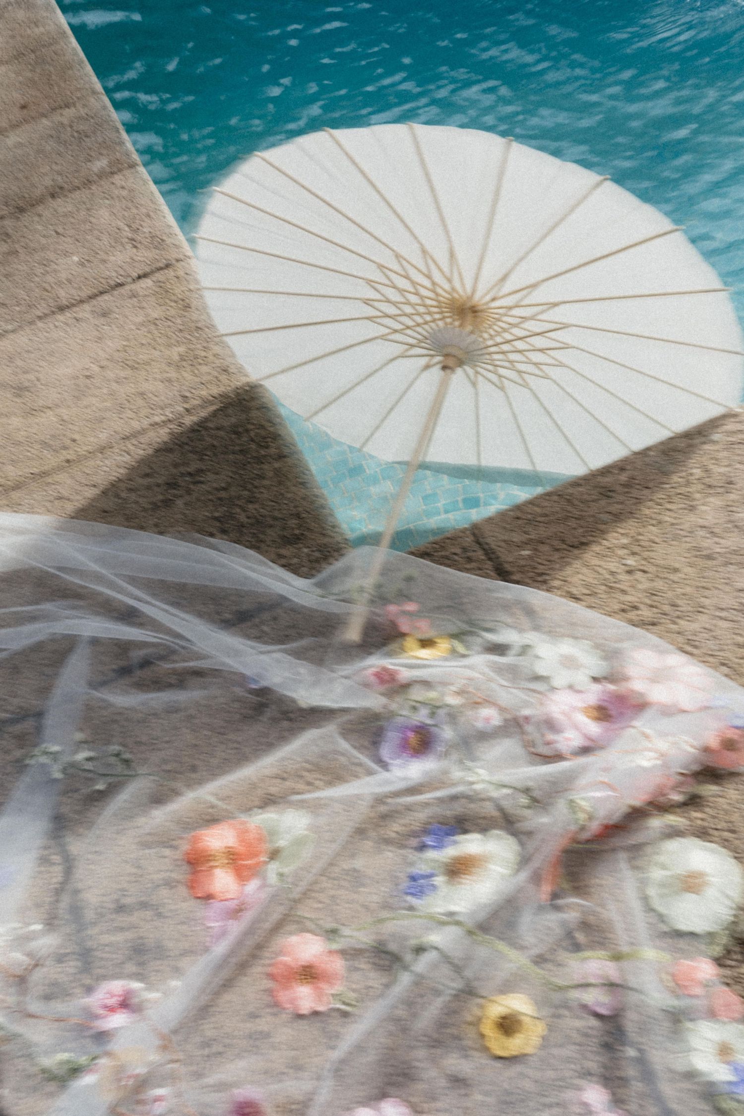White paper parasol beside a floral-embroidered wedding veil draped near a turquoise swimming pool edge.