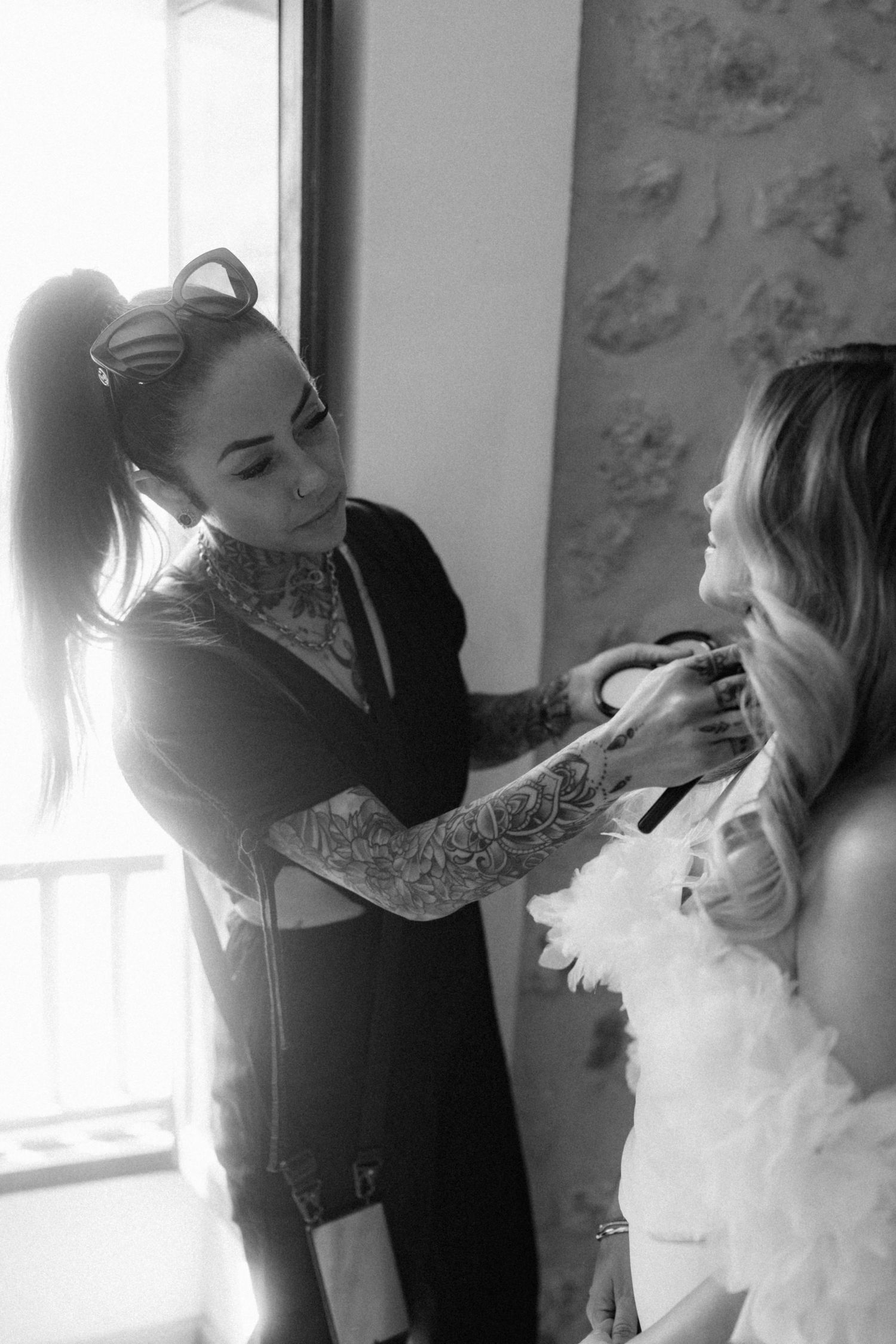 Heavily tattooed makeup artist applying makeup to a bride in a white feathered dress before her wedding.