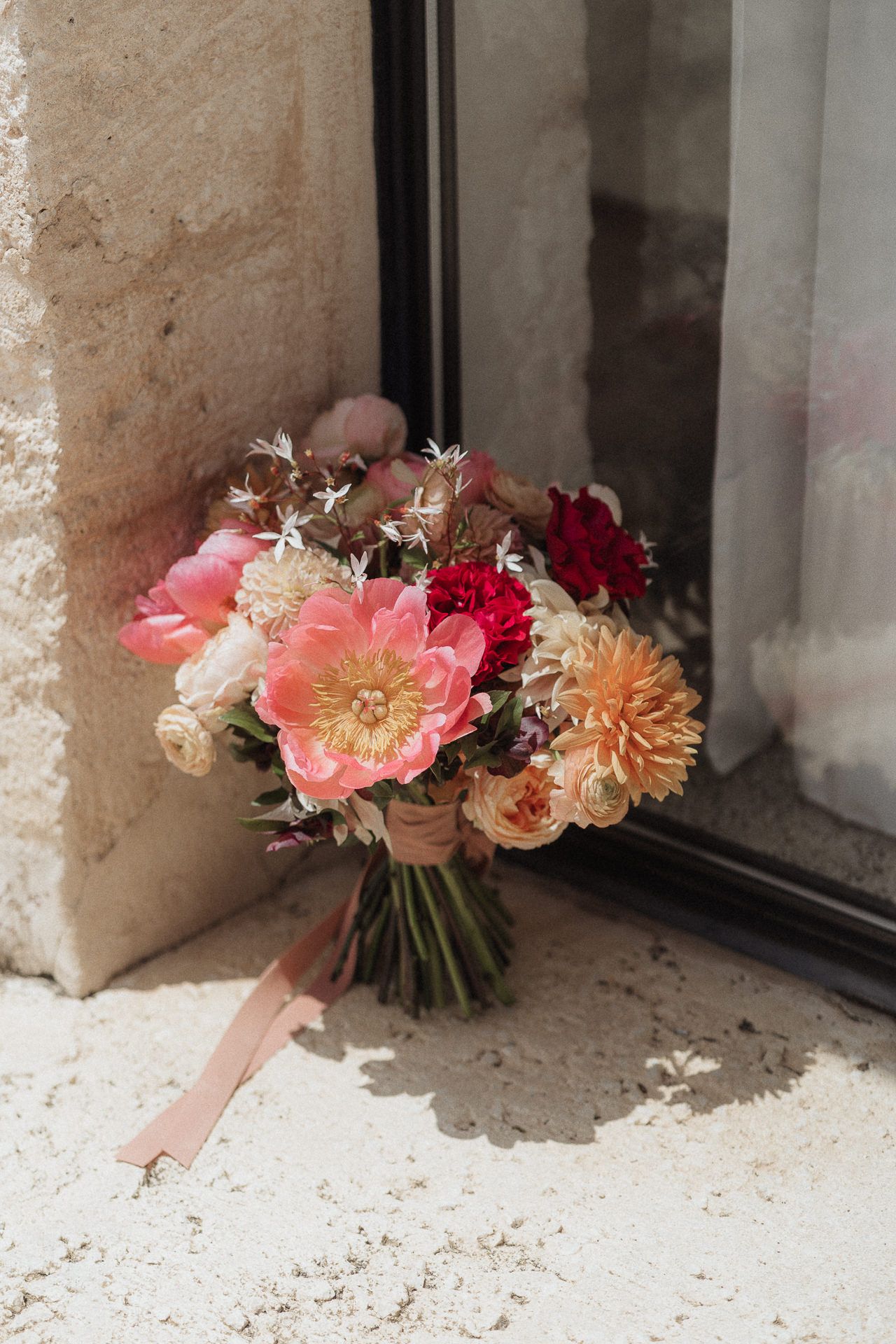Romantic bridal bouquet with pink peonies, coral dahlias, and red roses leaning against stone wall in natural light.
