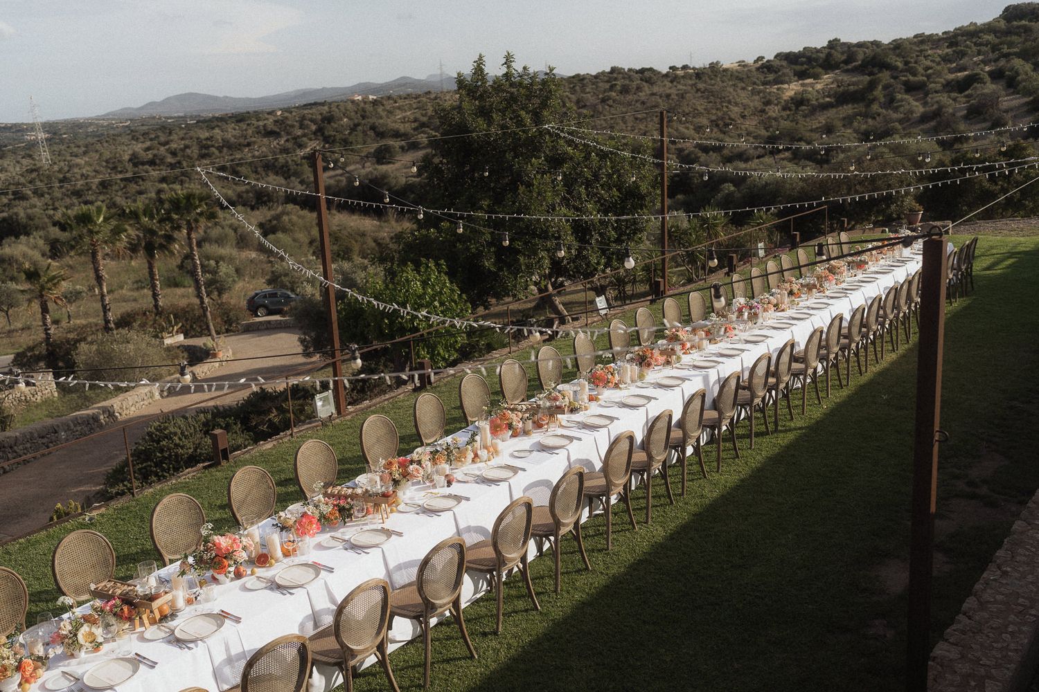 Elegant outdoor wedding reception with long white tables set for guests on a hillside venue with string lights.
