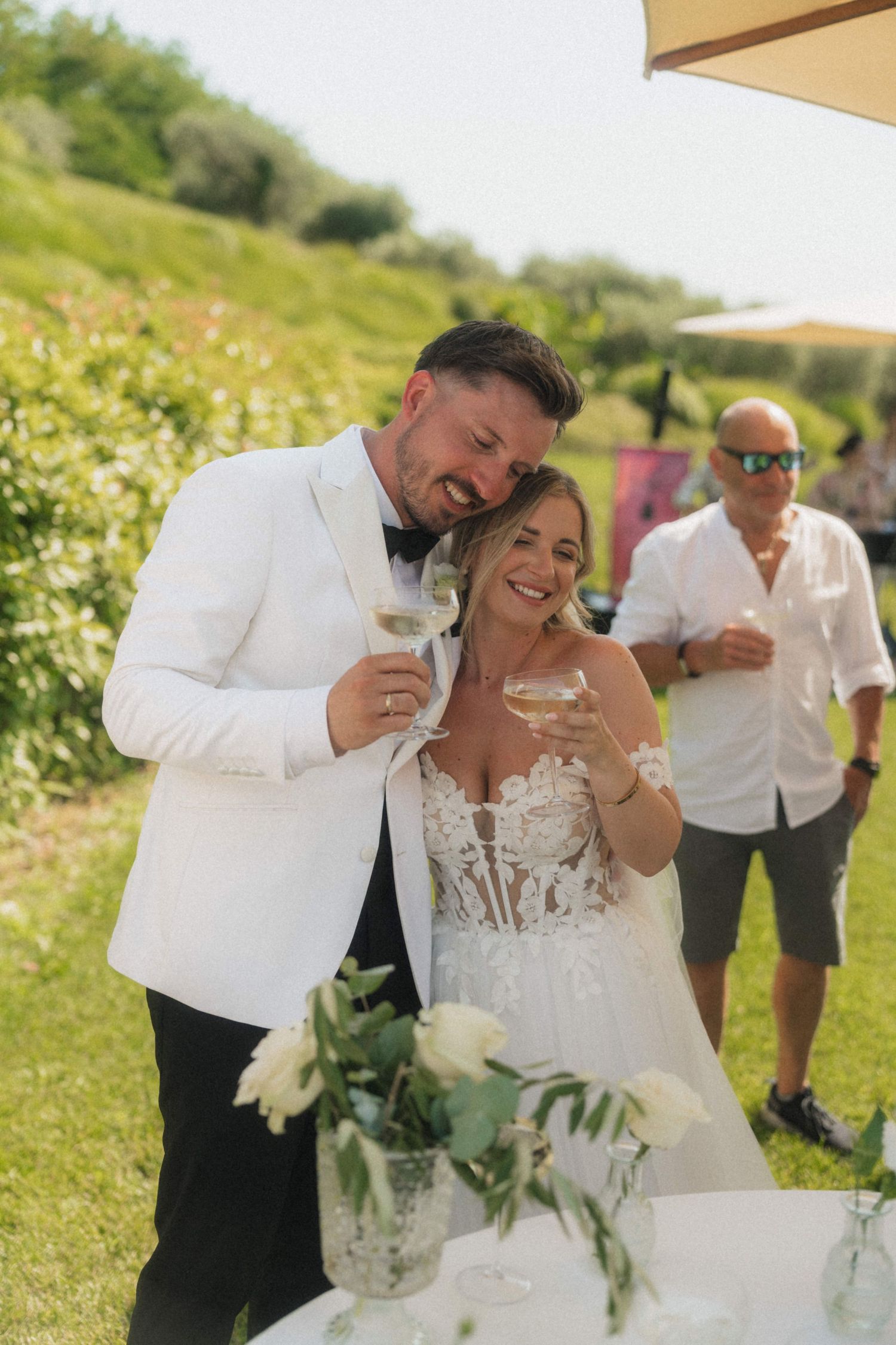 Bride and groom in white tuxedo toasting with champagne glasses at outdoor wedding reception.