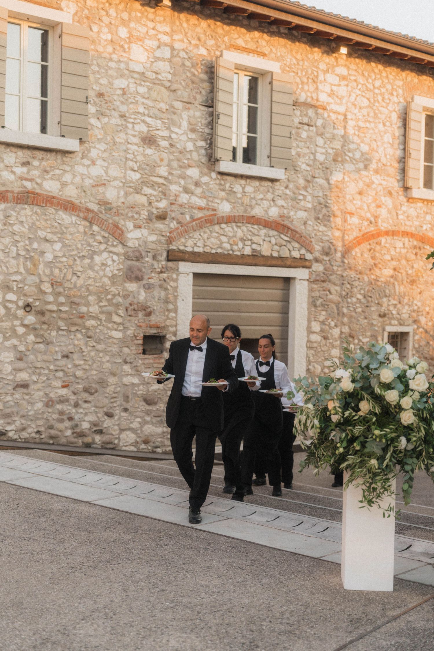 Three waiters in formal attire carry serving trays outside a rustic stone building at a wedding reception.