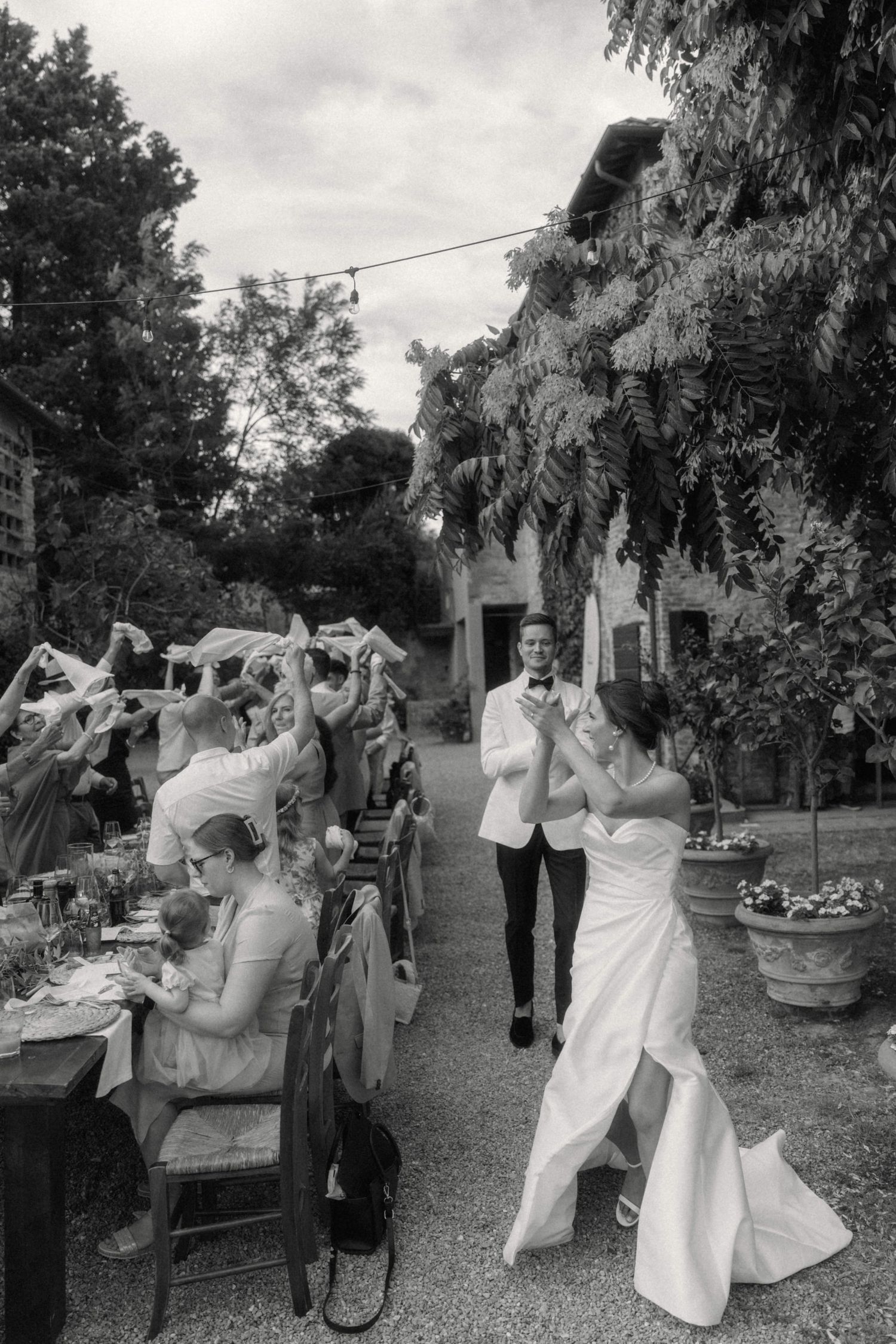 Black and white photo of bride and groom making a grand entrance to cheering guests at outdoor wedding reception.