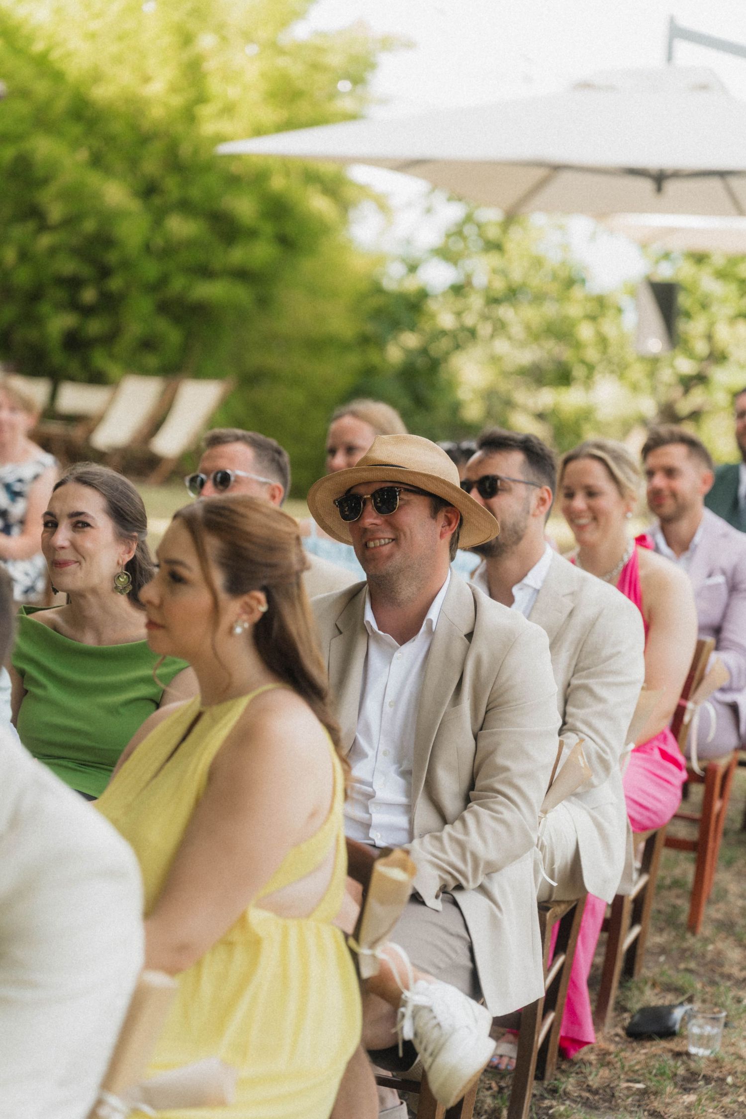 Wedding guests seated outdoors in summer attire watching ceremony under garden umbrella.