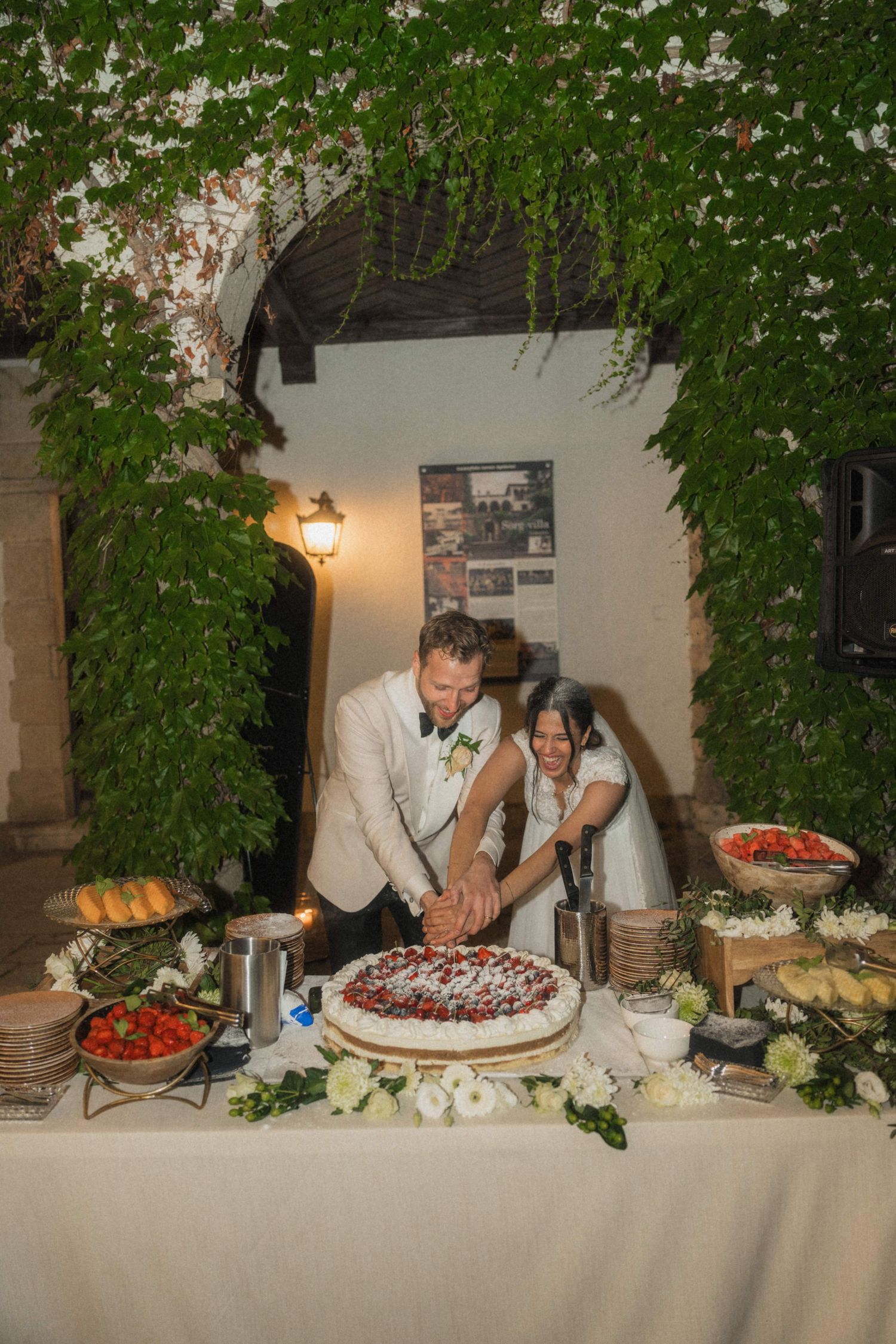 Bride and groom cutting wedding cake at night under lush green foliage at rustic venue.