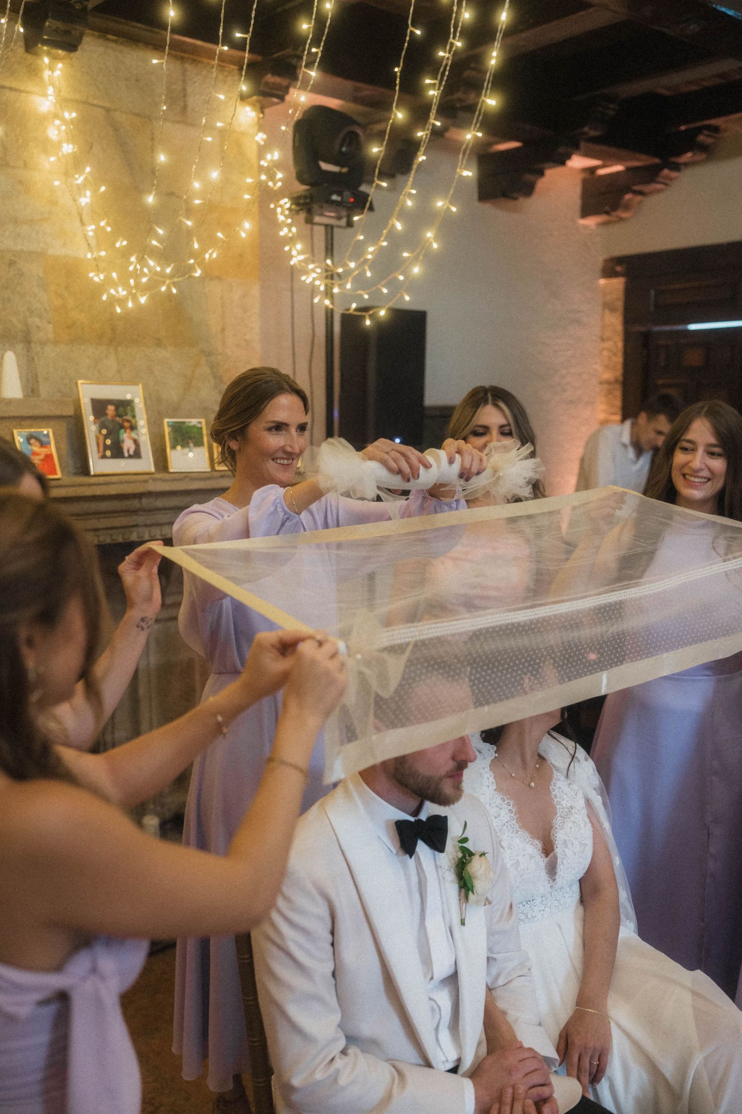 Bridesmaids lifting wedding veil over seated bride and groom at fairy light decorated indoor reception.