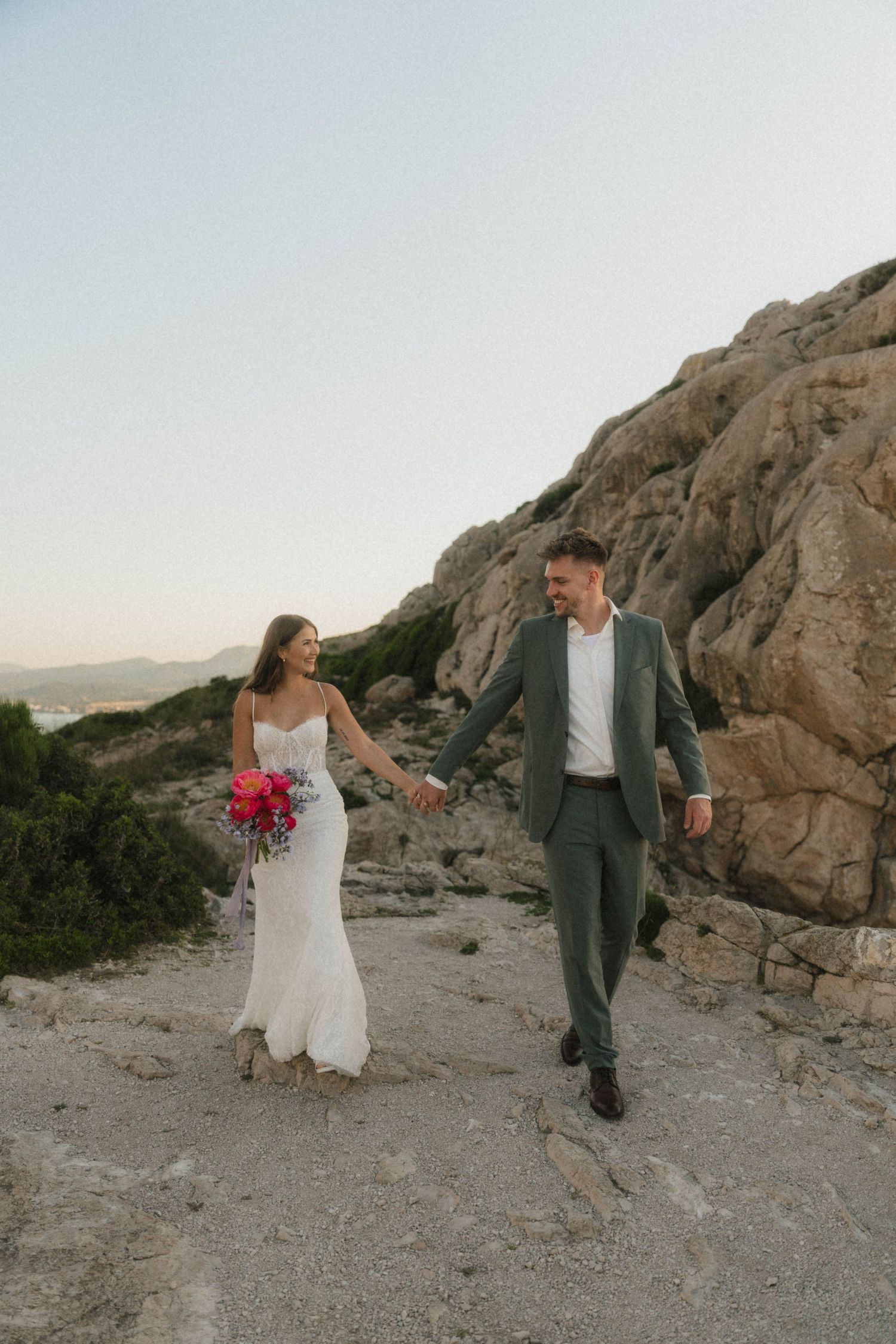 Bride and groom walk hand in hand along rocky coastal path, she in white gown holding pink bouquet, he in gray suit.