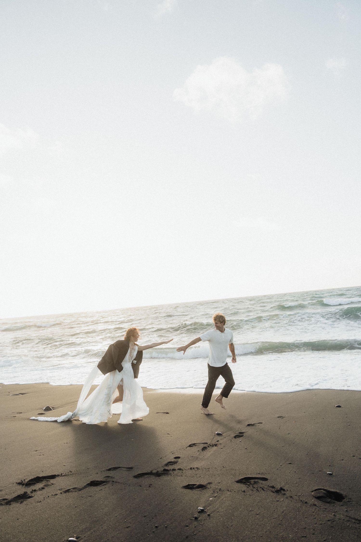 Joyful wedding couple holding hands running along black sand beach at the ocean shore.