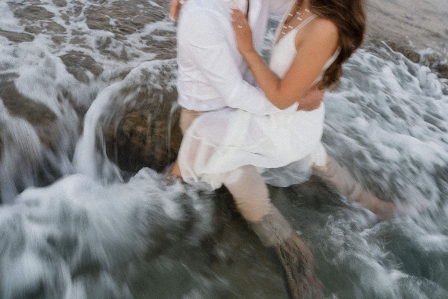 Couple embracing on rocks as ocean waves crash around them in white wedding outfits.
